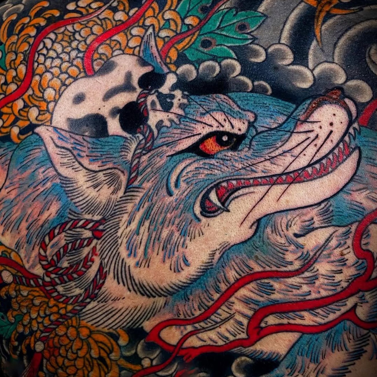 A colorful tattoo of a snarling wolf with a cartoonish style, featuring vibrant shades of blue, pink, and red, with a background of foliage and abstract elements.