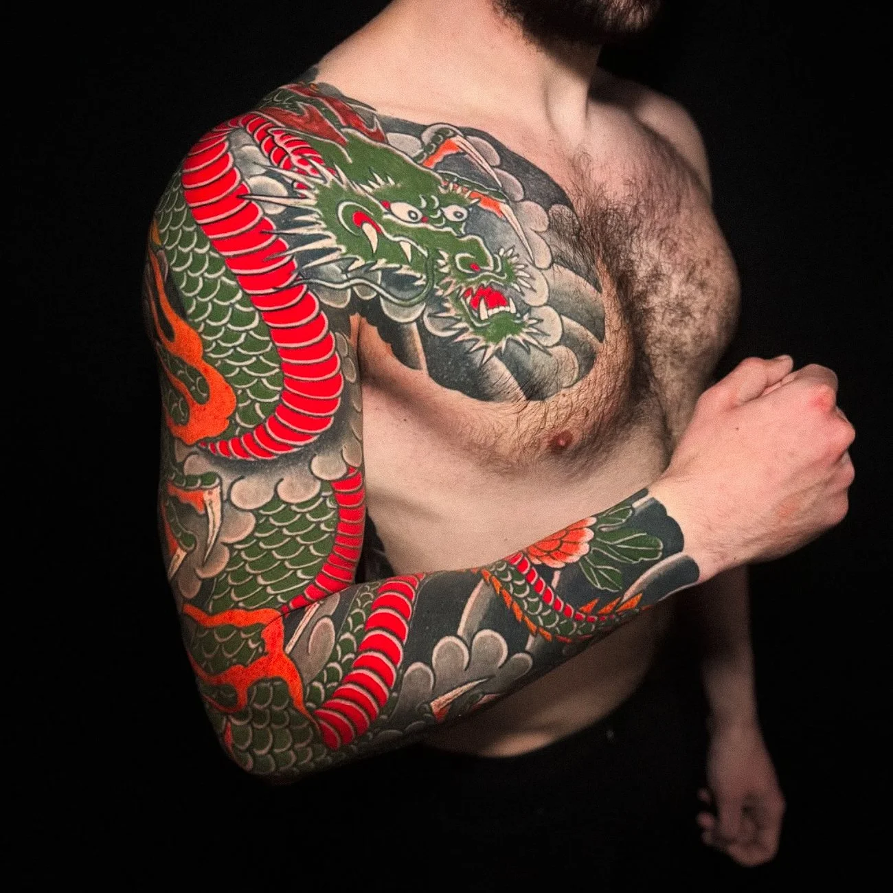 A shirtless man with a full sleeve tattoo of a colorful dragon on his left arm and part of his chest. The tattoo features green, red, orange, and black colors with traditional Asian design elements.