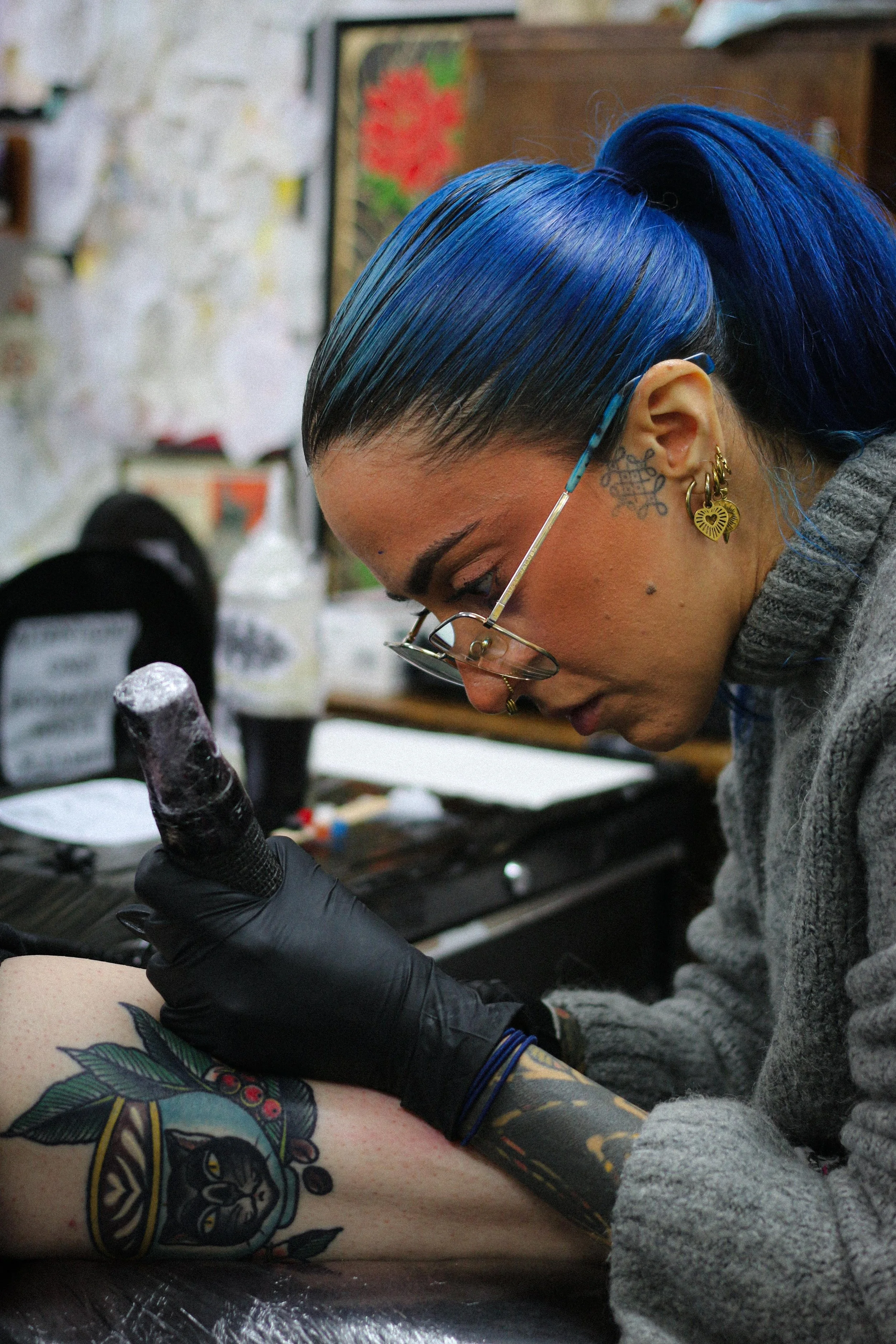 A woman with blue hair, glasses, and tattoos, wearing black gloves, is tattooing a colorful cat design on a person's arm in a tattoo parlor.