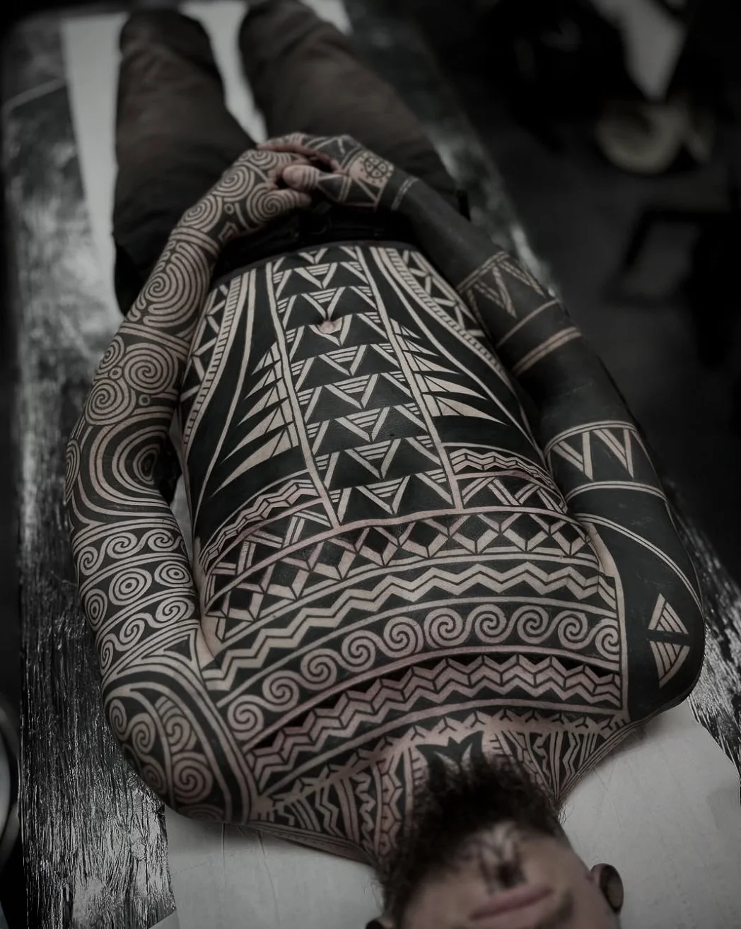 Person lying on their back with intricate black and white tribal tattoos covering their arms, chest, and stomach, while holding their hands on their lower abdomen.
