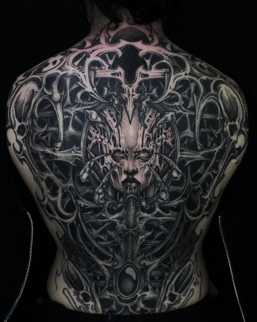 Black and gray intricate tattoo covering a person's back, featuring a central human face with closed eyes, mechanical and organic elements, and abstract designs.