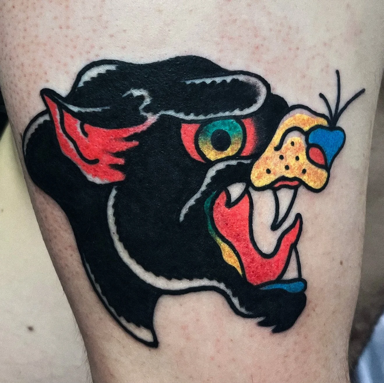 Tattoo of a stylized lion with its mouth open, featuring a colorful butterfly and a leaf near its eye, with black and red colors.