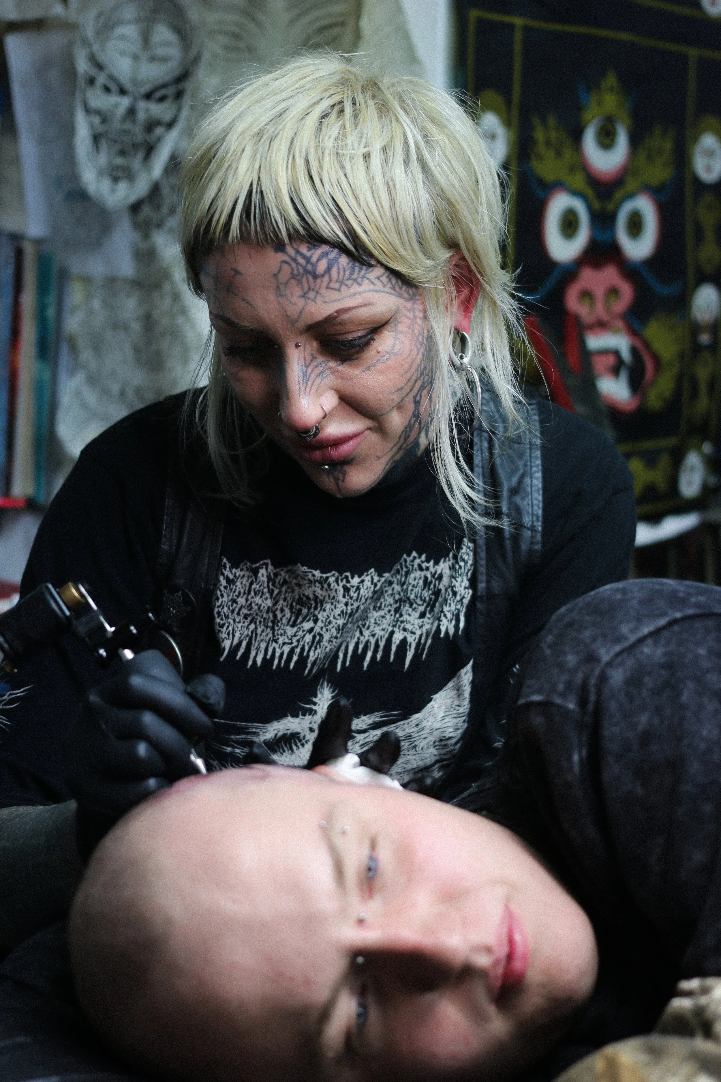 A tattoo artist with blonde hair and multiple facial piercings is tattooing a person's forehead in a studio decorated with artwork and paintings.
