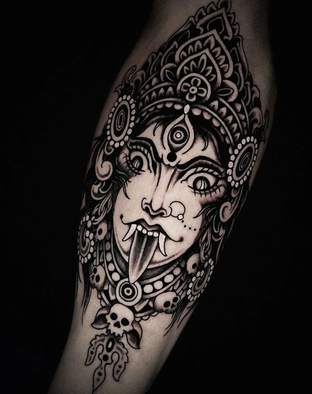 Black and gray tattoo of a fierce woman with fangs and a tongue, wearing an elaborate headdress with skulls and beads, on a person's arm.