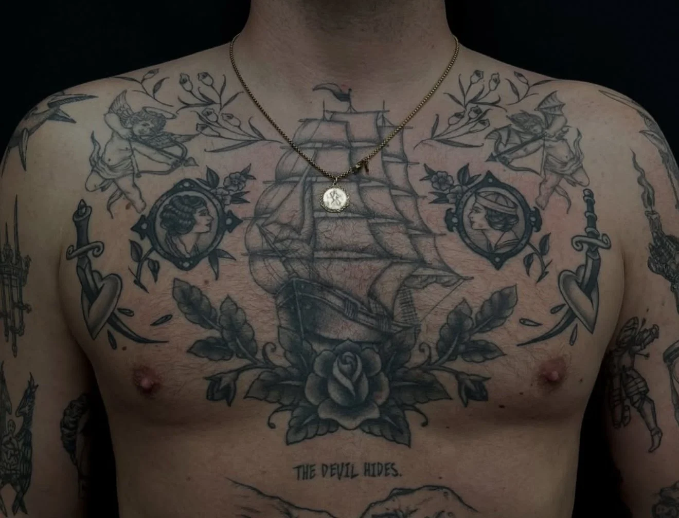 Black and gray chest tattoos featuring a sailing ship, portraits of women, angels, a sword, roses, and the phrase "The devil hides." The person is wearing a necklace with a coin pendant.
