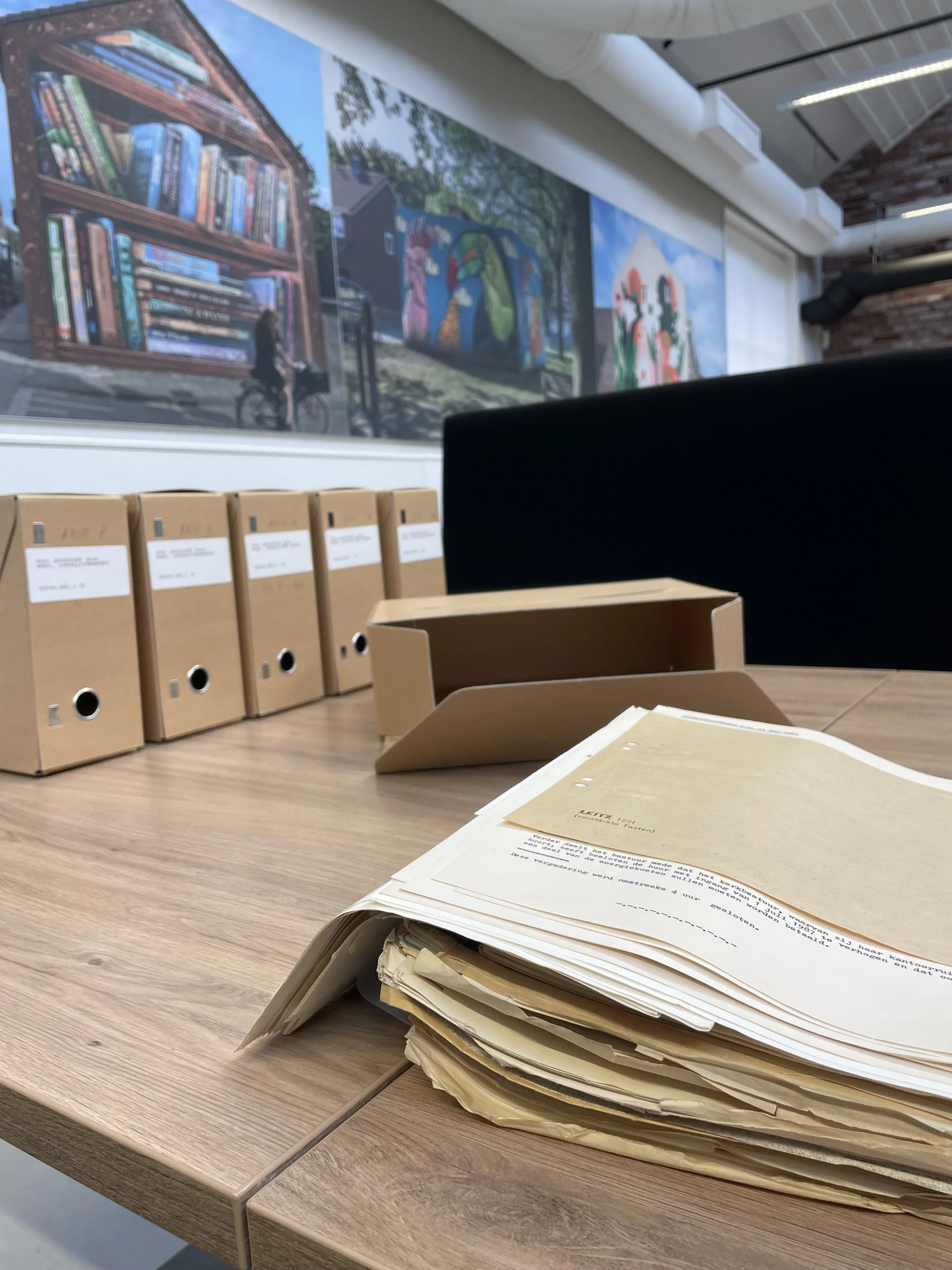 A table with stacks of papers and manila folders in an office or study area. Large colorful murals of bookshelves, outdoor scenes, and artwork are visible on the wall in the background.