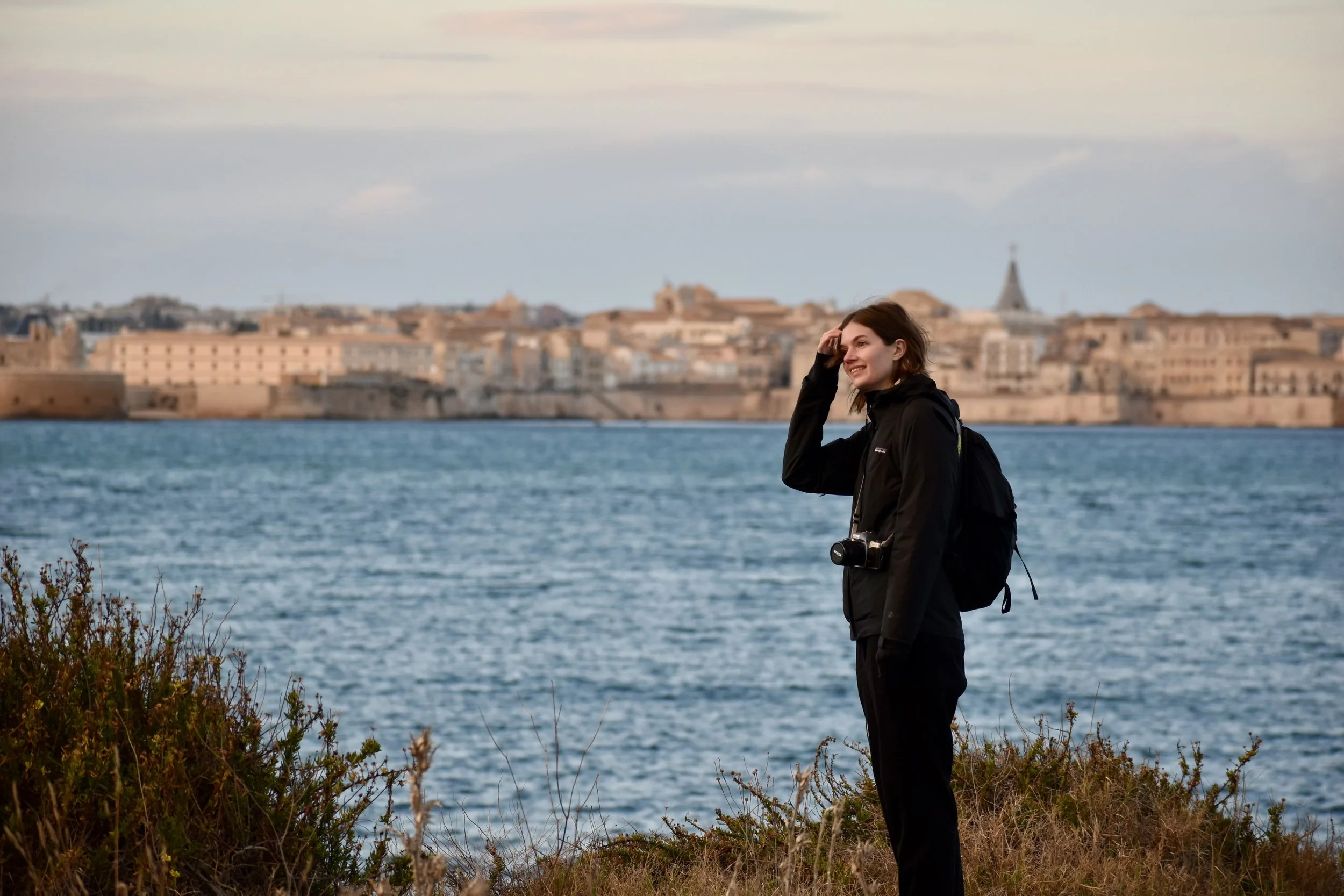 Stephanie (a white woman with mid-long brown wavy hair), wearing all black and carrying a camera. She is standing on a cliff top near the ocean, looking into the distance. In the background a blue ocean and the city Syracuse are visible.