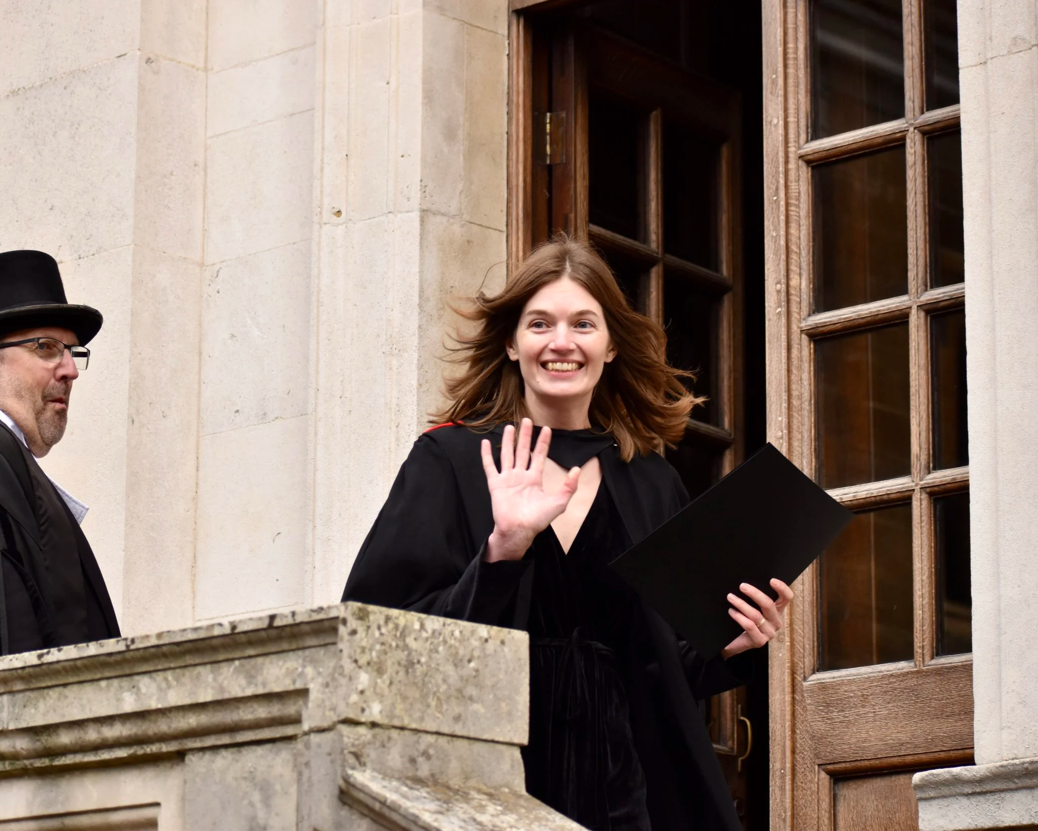A young woman in a black graduation gown waving and smiling as she steps outside a building, holding a diploma.