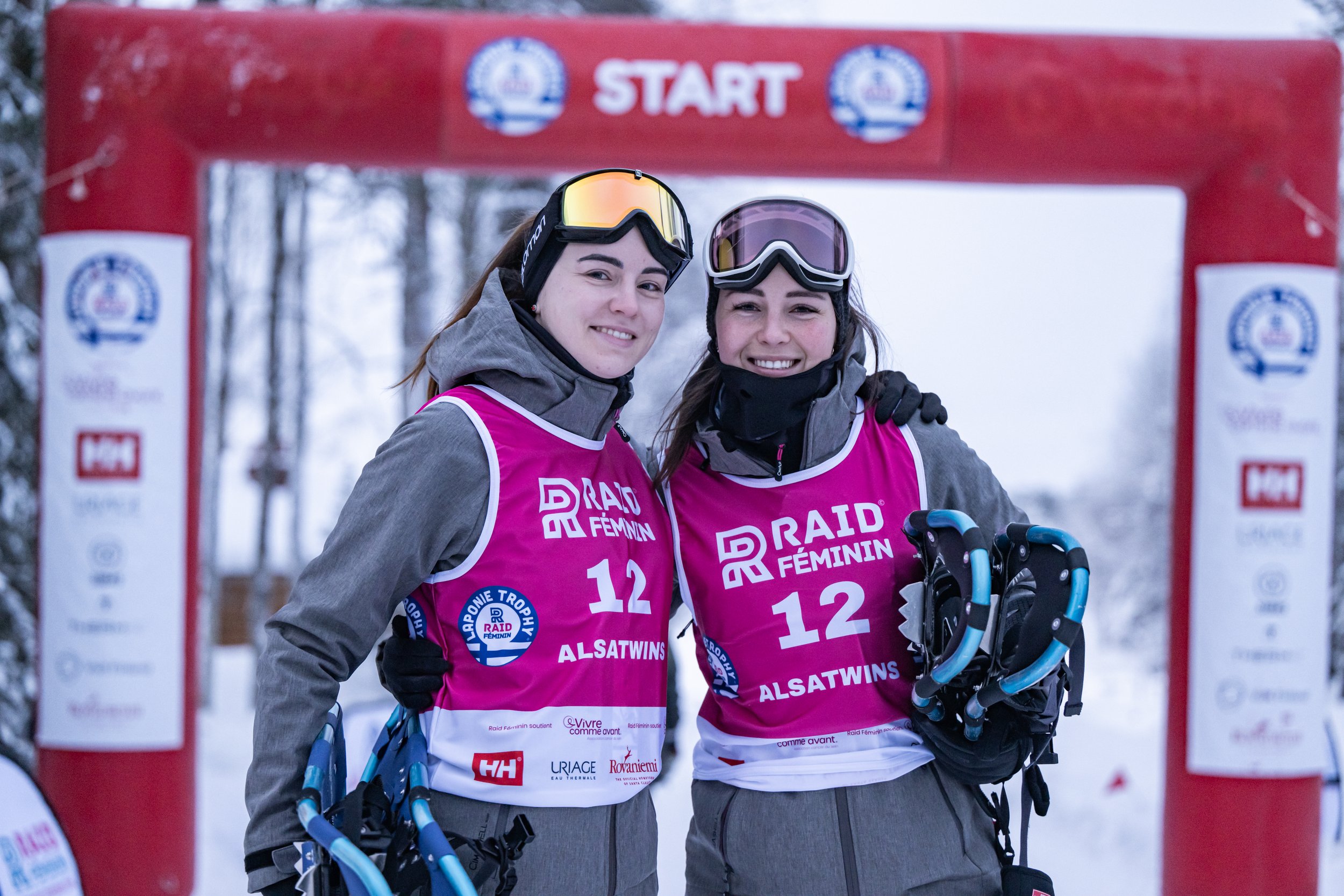 Deux femmes en ski alpin, portant des vestes gris et des gilets roses avec le logo 'Raid Féminin'. Elles sourient, se tiennent sous une arche de départ rouge avec la neige et des arbres en arrière-plan.