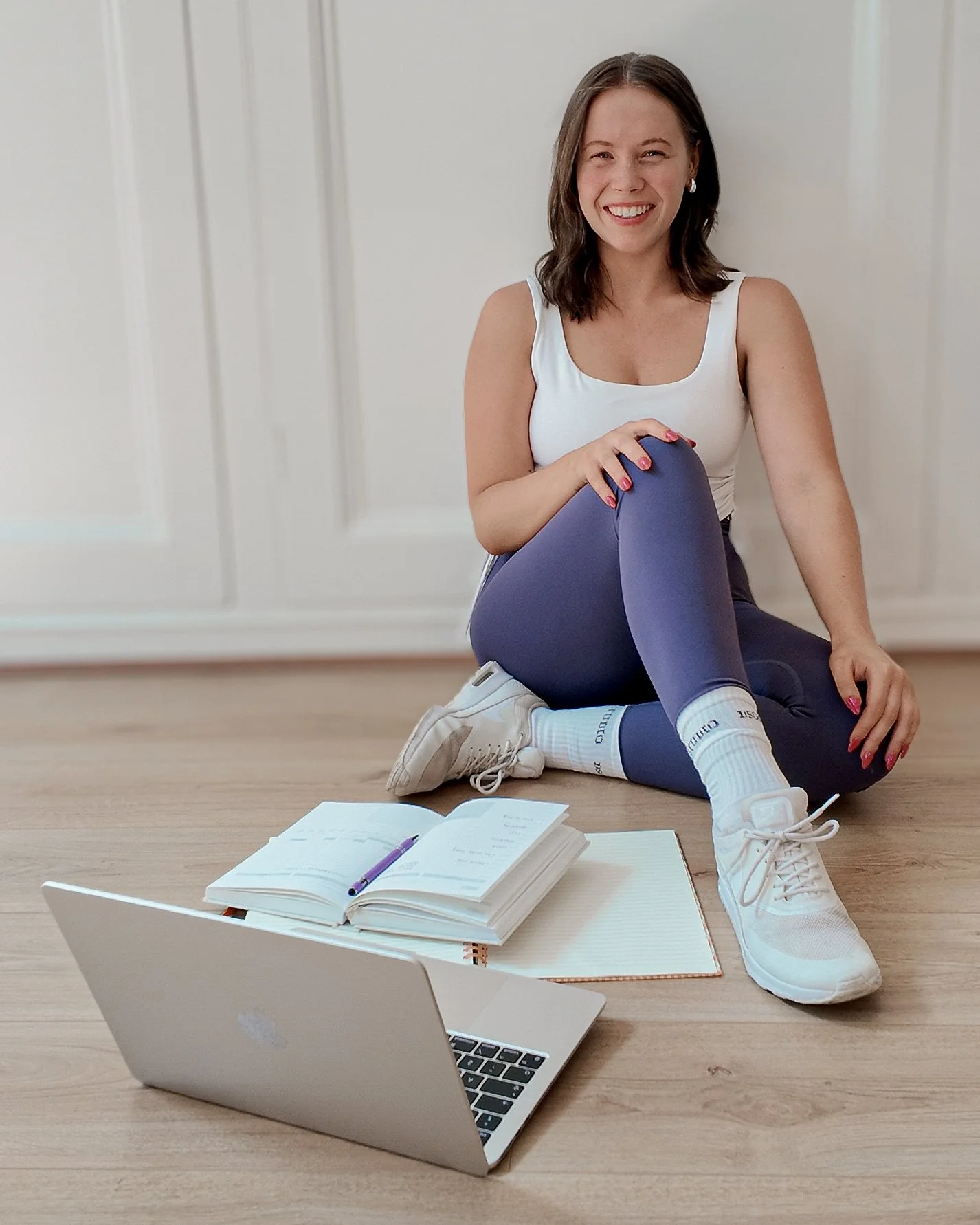 A young woman sitting on the floor with a laptop, open notebook, and pen in front of her, smiling and looking at the camera. She is wearing athletic clothes including a white tank top, purple leggings, and white sneakers.