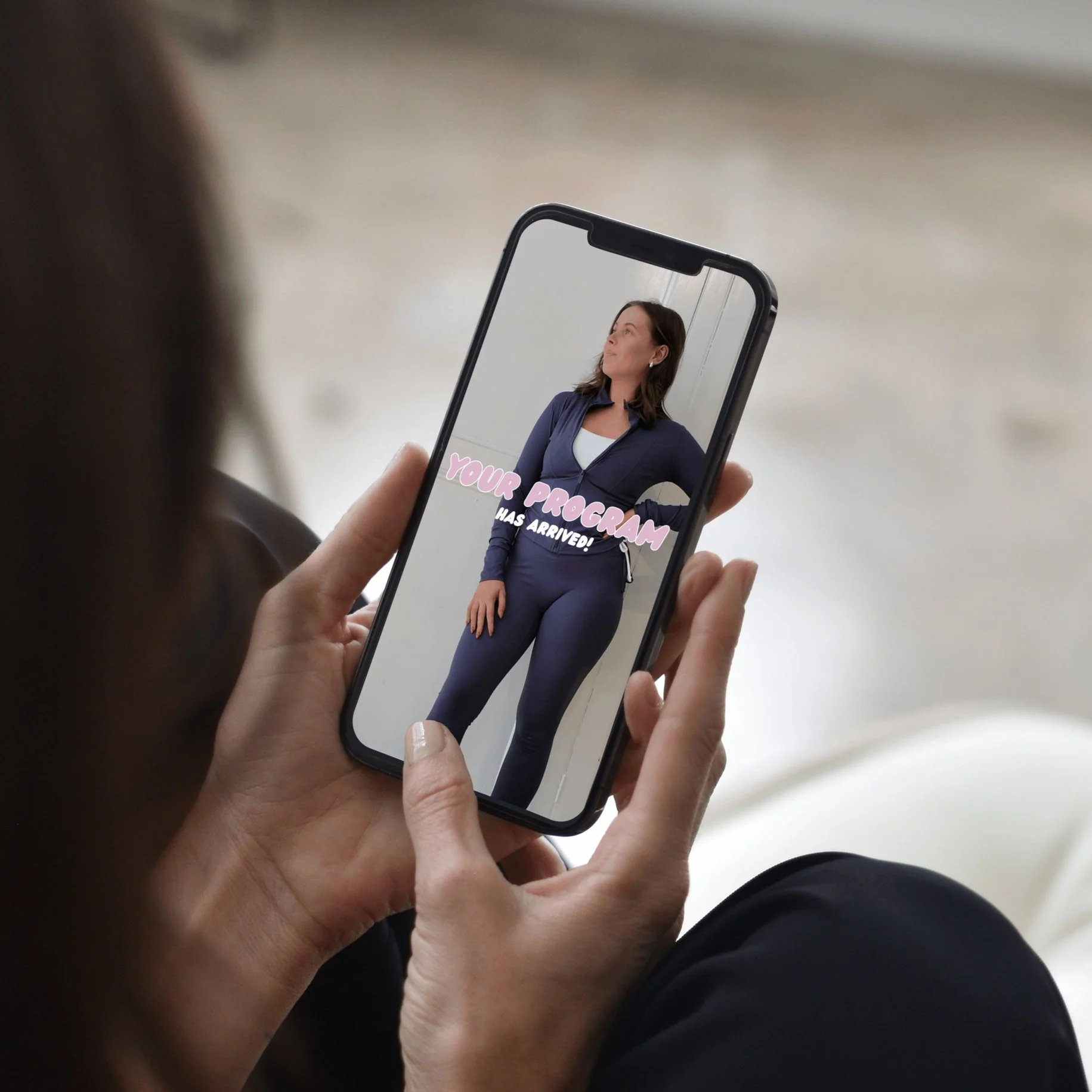 Person holding a smartphone displaying a photo of a woman in workout clothes with text overlay that says 'Your Program Has Arrived!'
