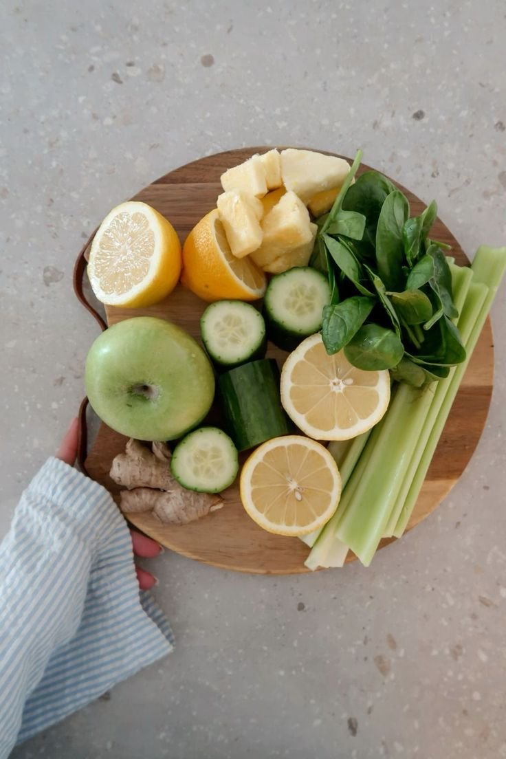 A wooden round cutting board with lemon, pineapple, apple, cucumber, ginger, and spinach arranged for preparing a healthy meal or juice.