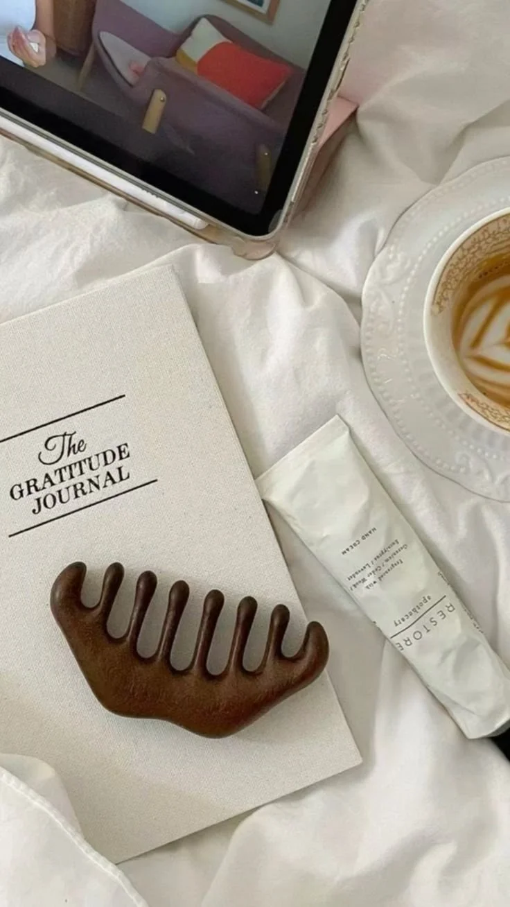 Tablet displaying a colorful couch image, white gratitude journal, chocolate reindeer-shaped rest relaxation tool, sachet of Les Cinq Filles skincare product, and a cup of coffee with swirled cream on a white bed sheet.