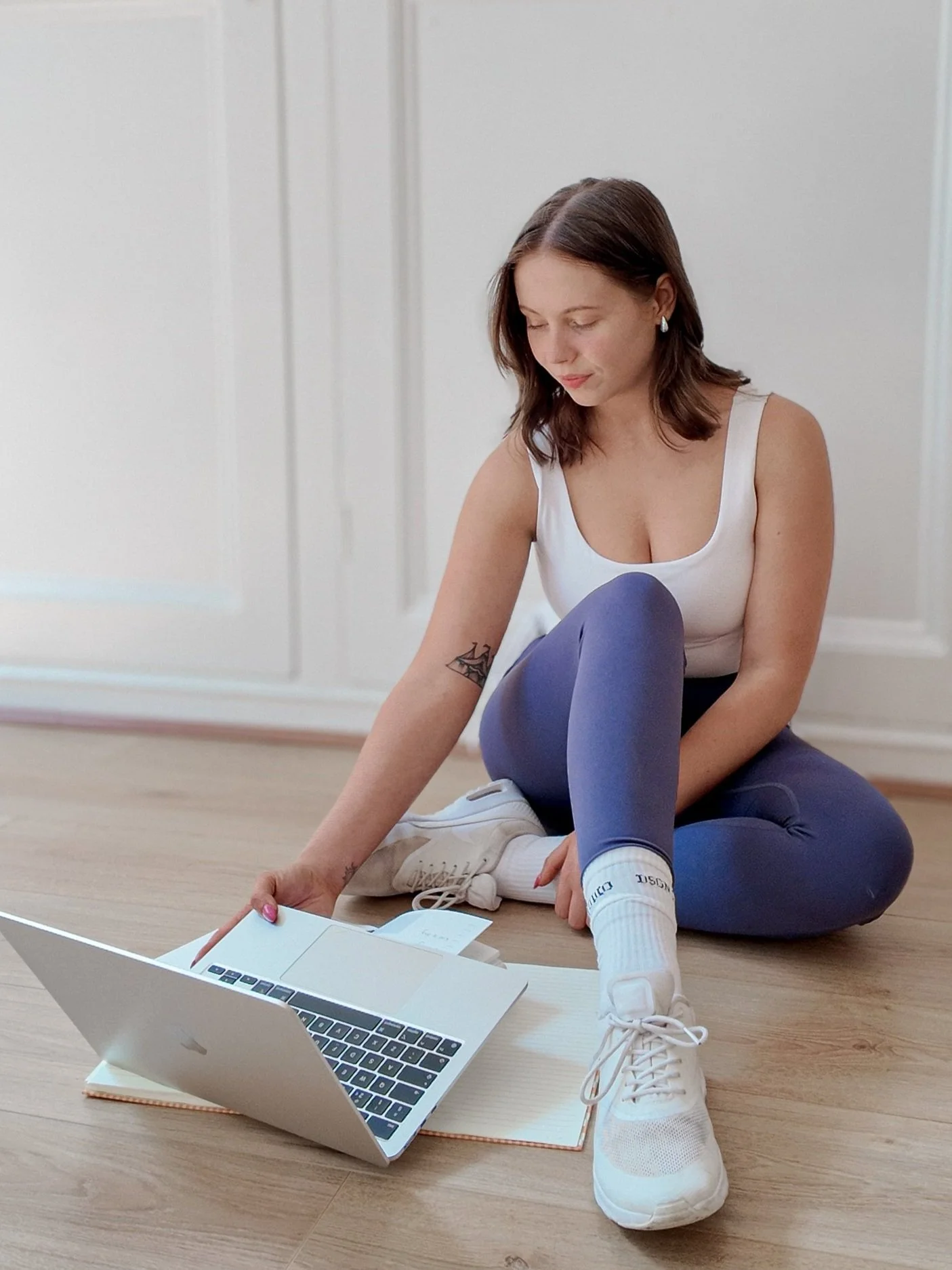 A woman sitting on the floor with a laptop, wearing athletic clothing and white sneakers, in a room with wooden flooring and white walls.