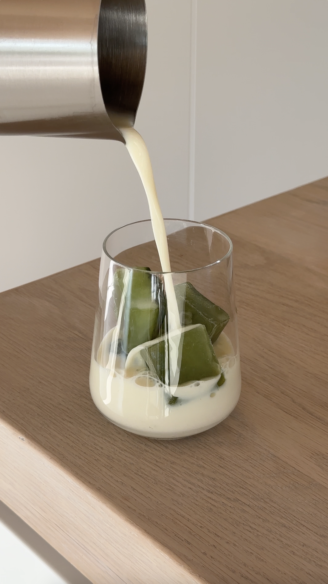Green jelly cubes in a glass with a creamy white drink being poured over them from a metal pitcher, on a wooden surface.