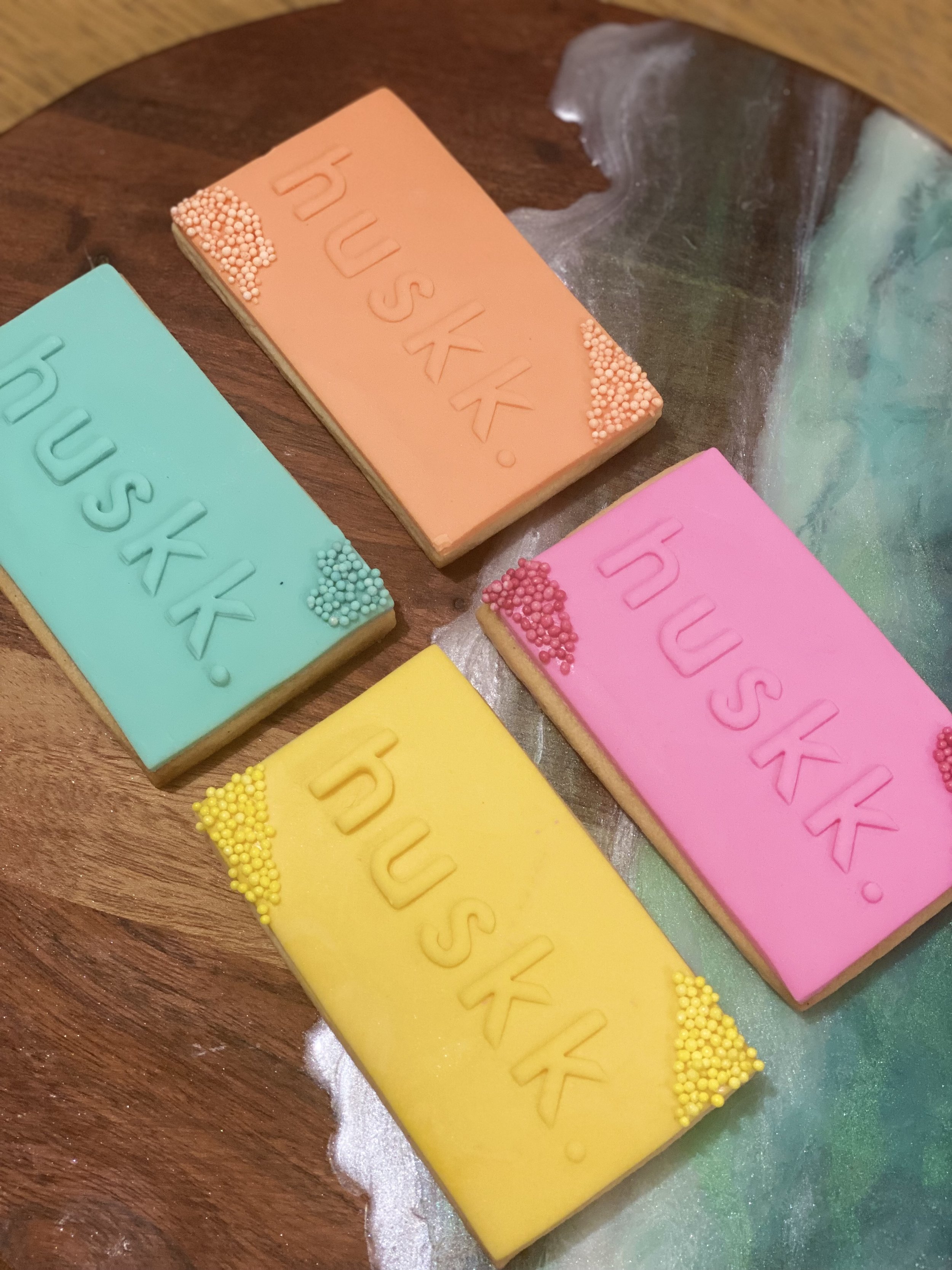 Four rectangular cookies decorated with pastel-colored icing and colorful sprinkles, arranged on a wooden surface. The cookies have the word "huskk" embossed in the icing.