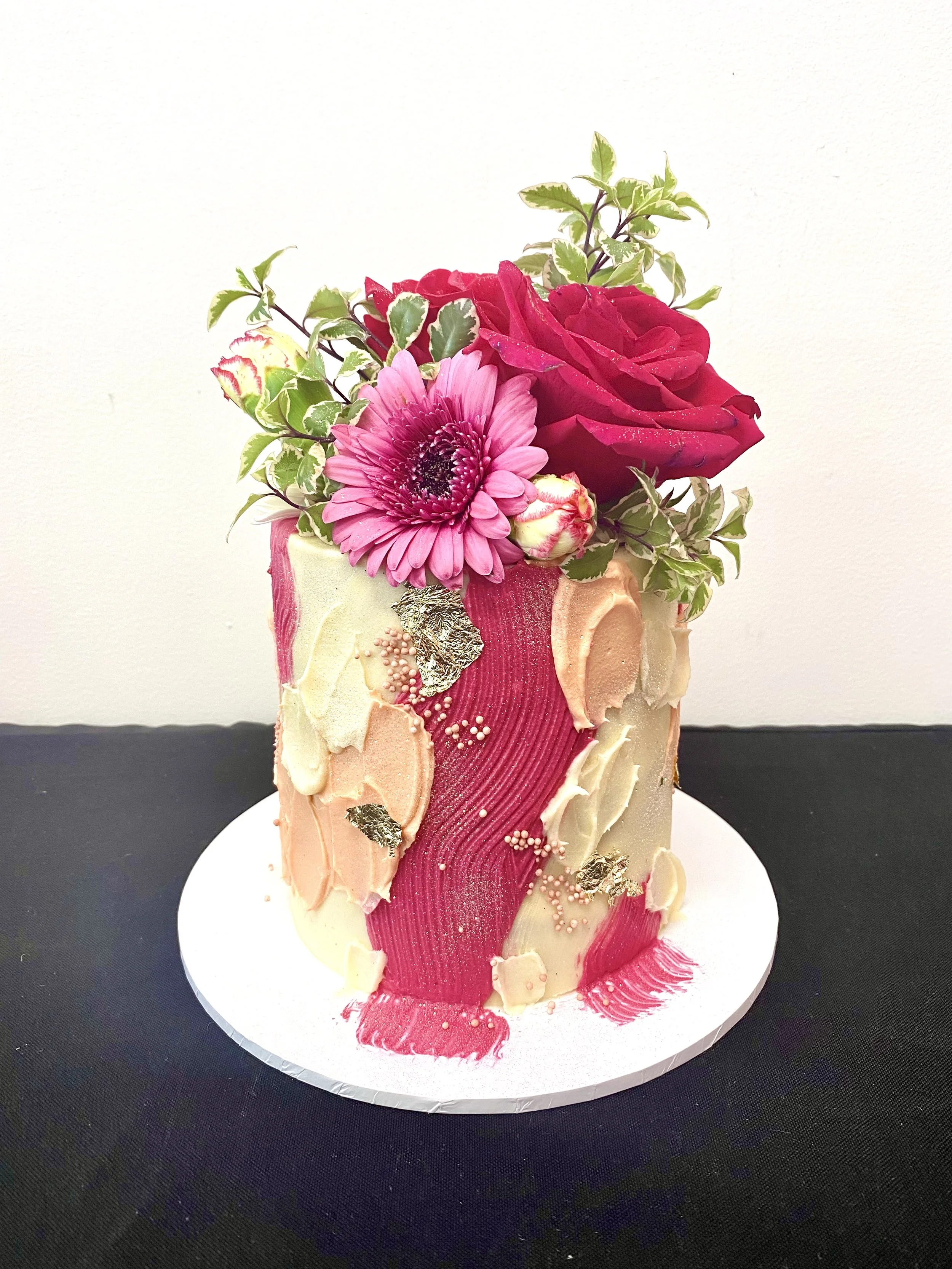 A floral cake with pink, white, and cream-colored frosting, topped with fresh roses, a pink gerbera daisy, and greenery.
