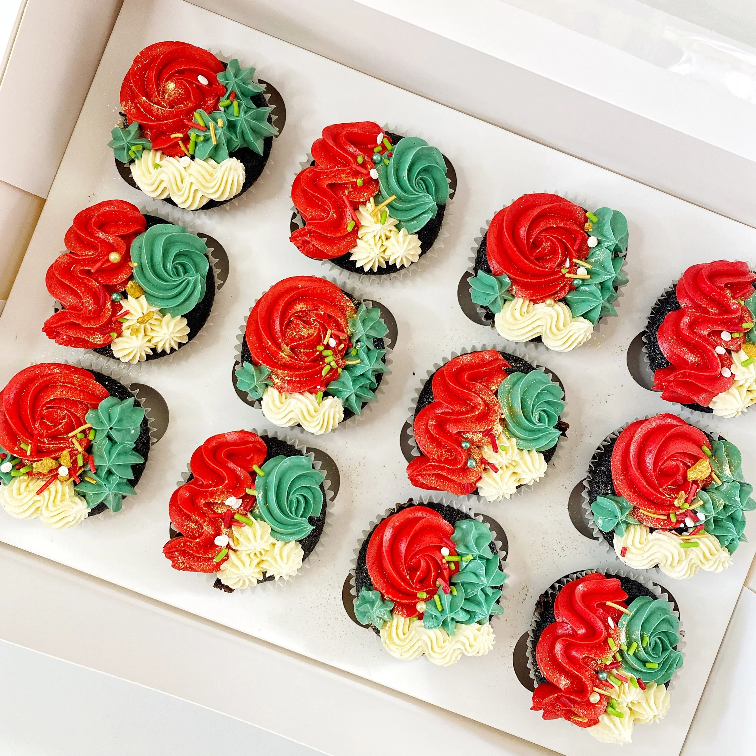 A box of decorated cupcakes with red, green, and white icing, topped with sprinkles and edible glitter.