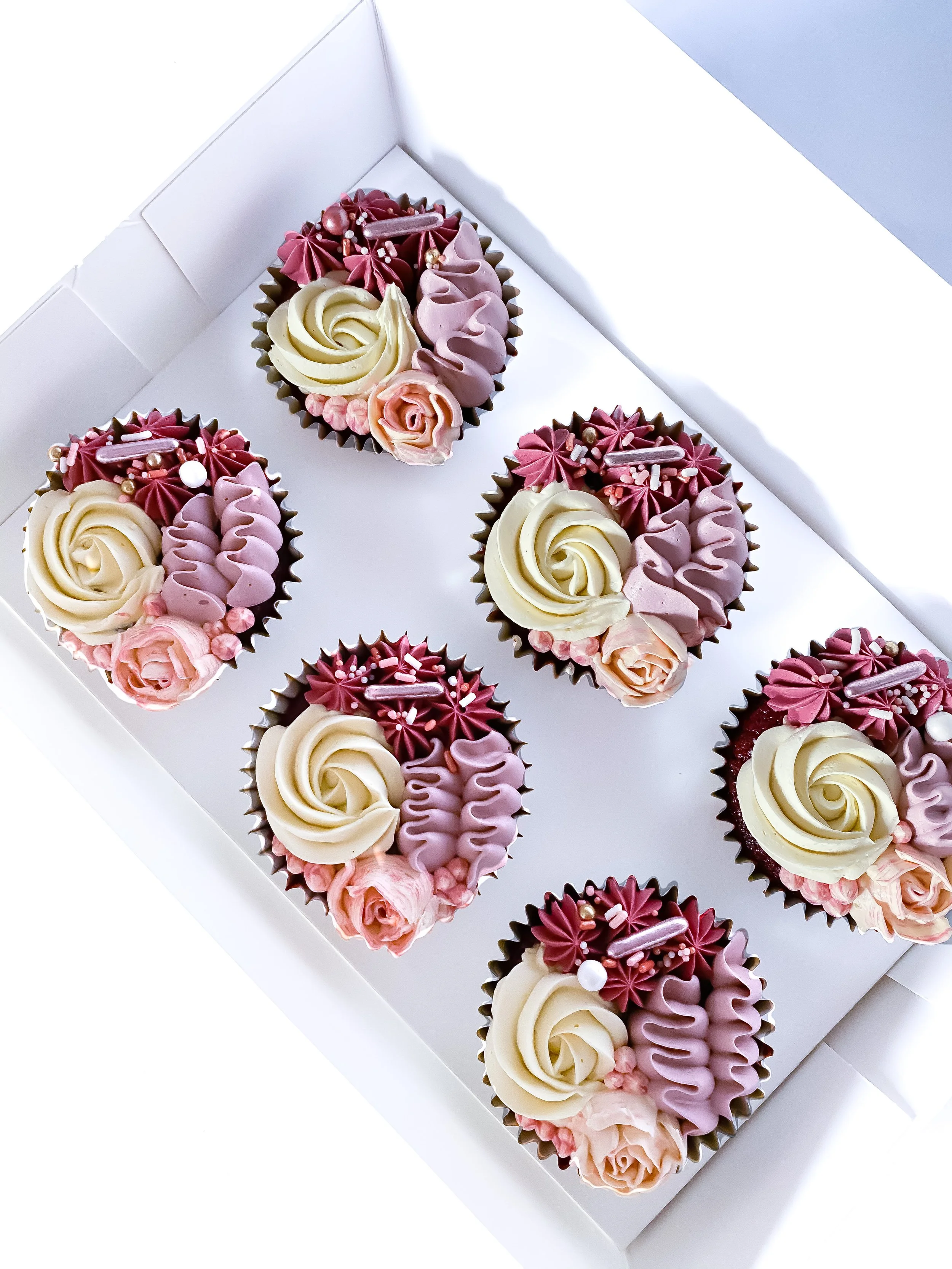 Six decorated cupcakes with pink, white, and purple frosting, sprinkles, and decorative flowers, arranged in a white box.