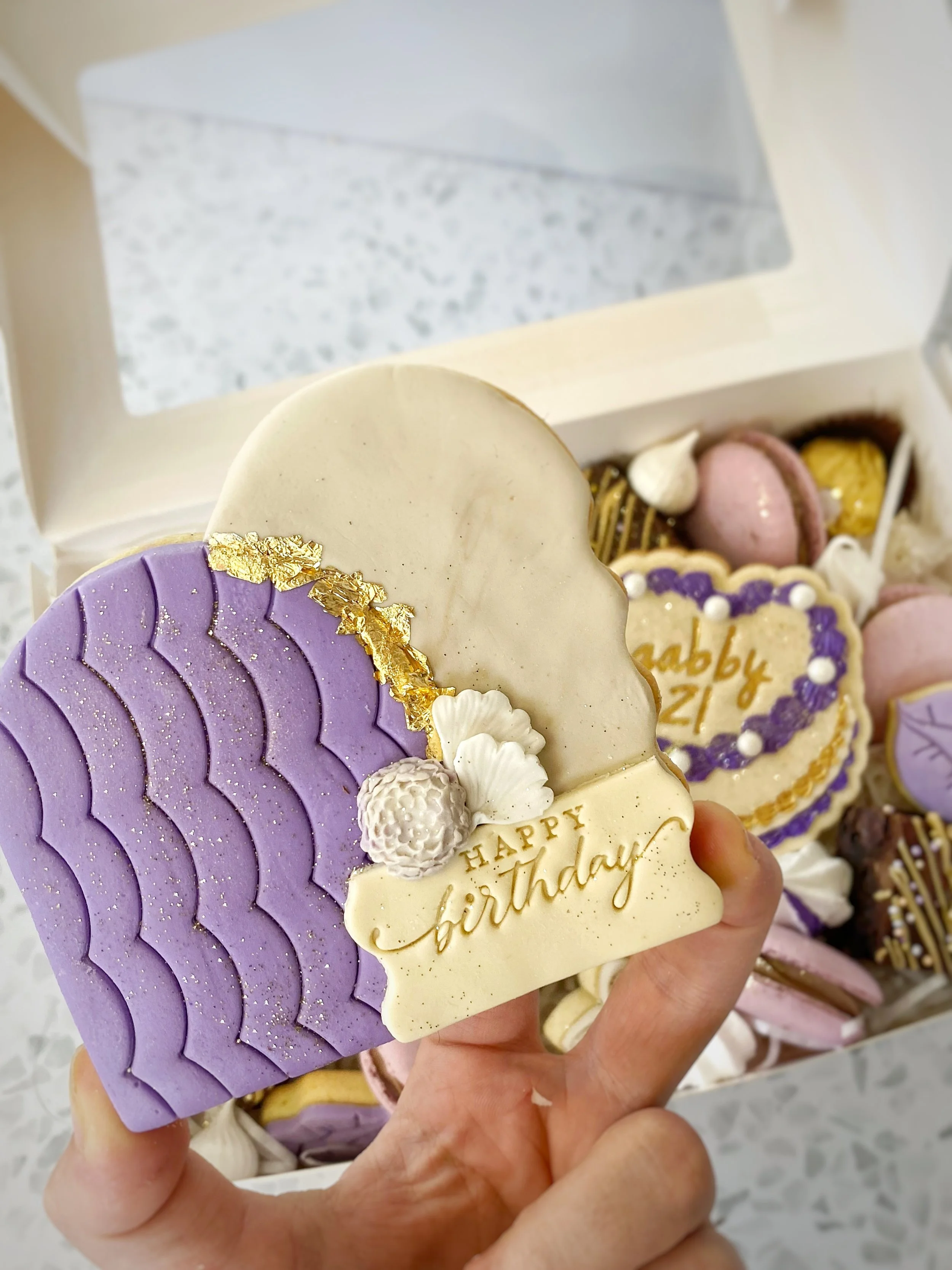 A hand holding a decorative birthday cookie with purple, white, and gold accents, featuring a 'Happy Birthday' message, in front of a tray of assorted cookies and treats.