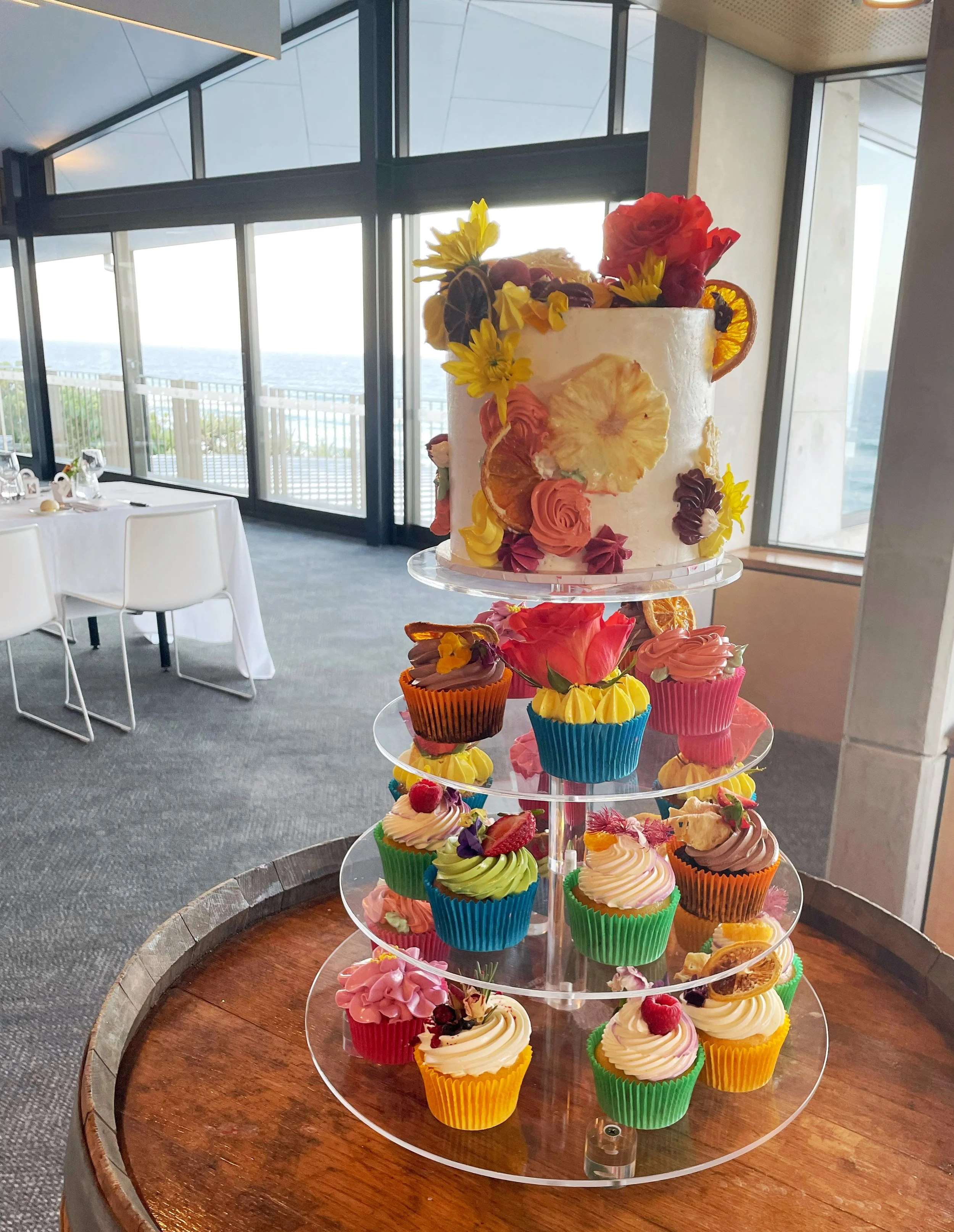 Colorful cupcake display with a large floral-decorated cake on top, set on a wooden table in a bright room with floor-to-ceiling windows and a view of the ocean.