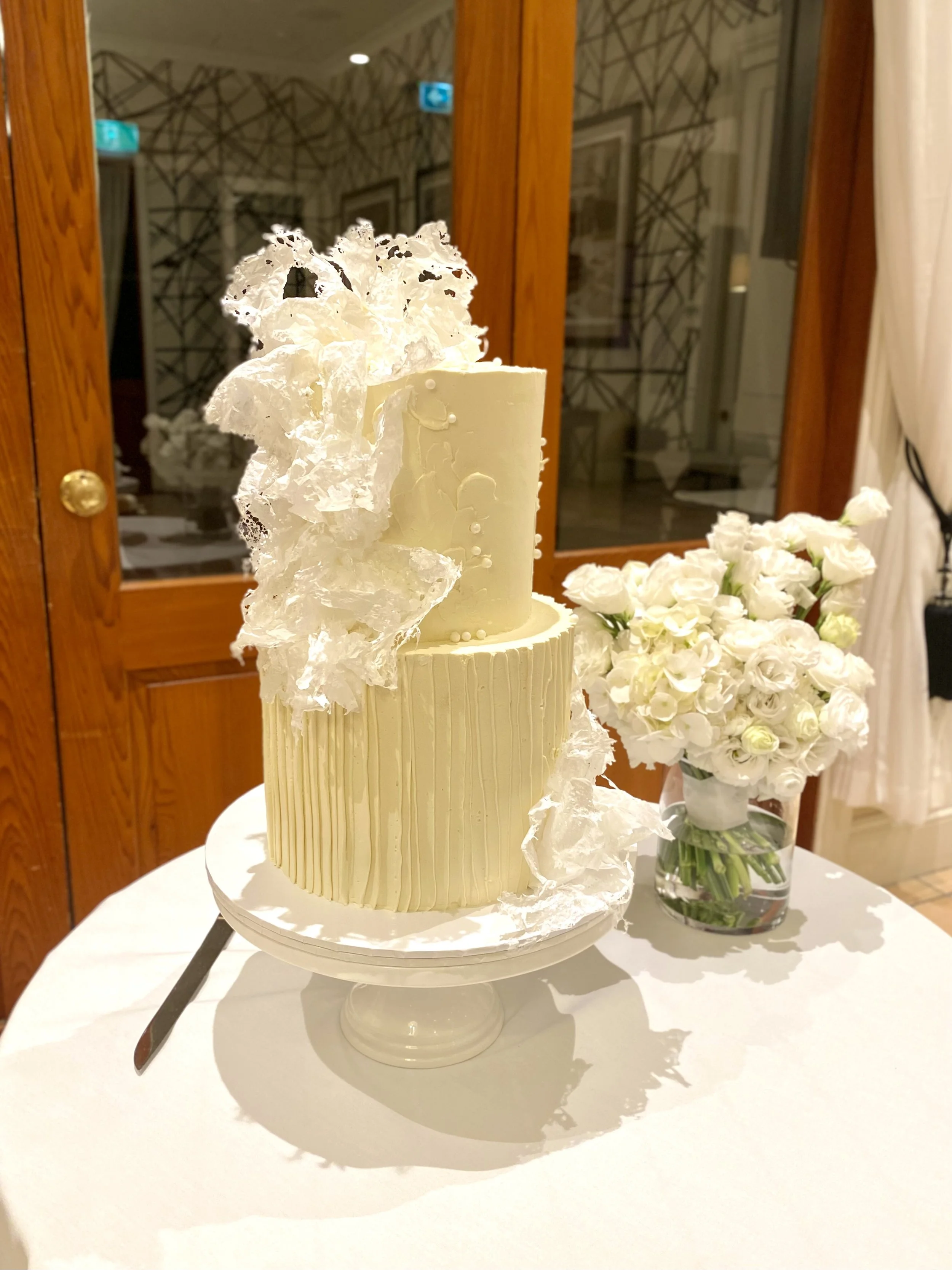 A two-tier white wedding cake with textured and smooth frosting, decorated with translucent sugar-like embellishments, placed on a pedestal stand. A bouquet of white flowers in a glass vase sits beside it on a white tablecloth.