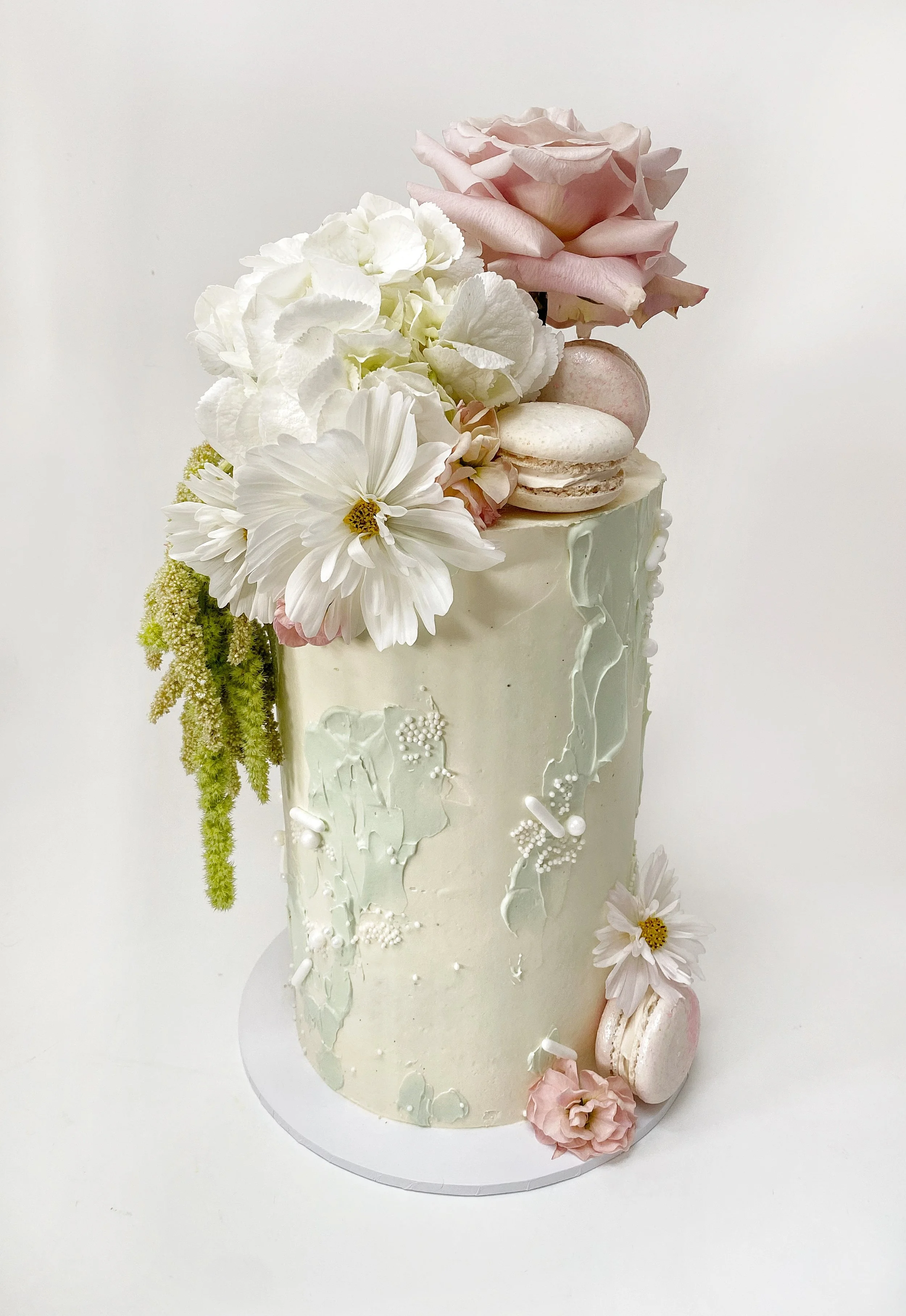 A tall white wedding cake decorated with white and pink flowers, macarons, and white sprinkles on a white background.