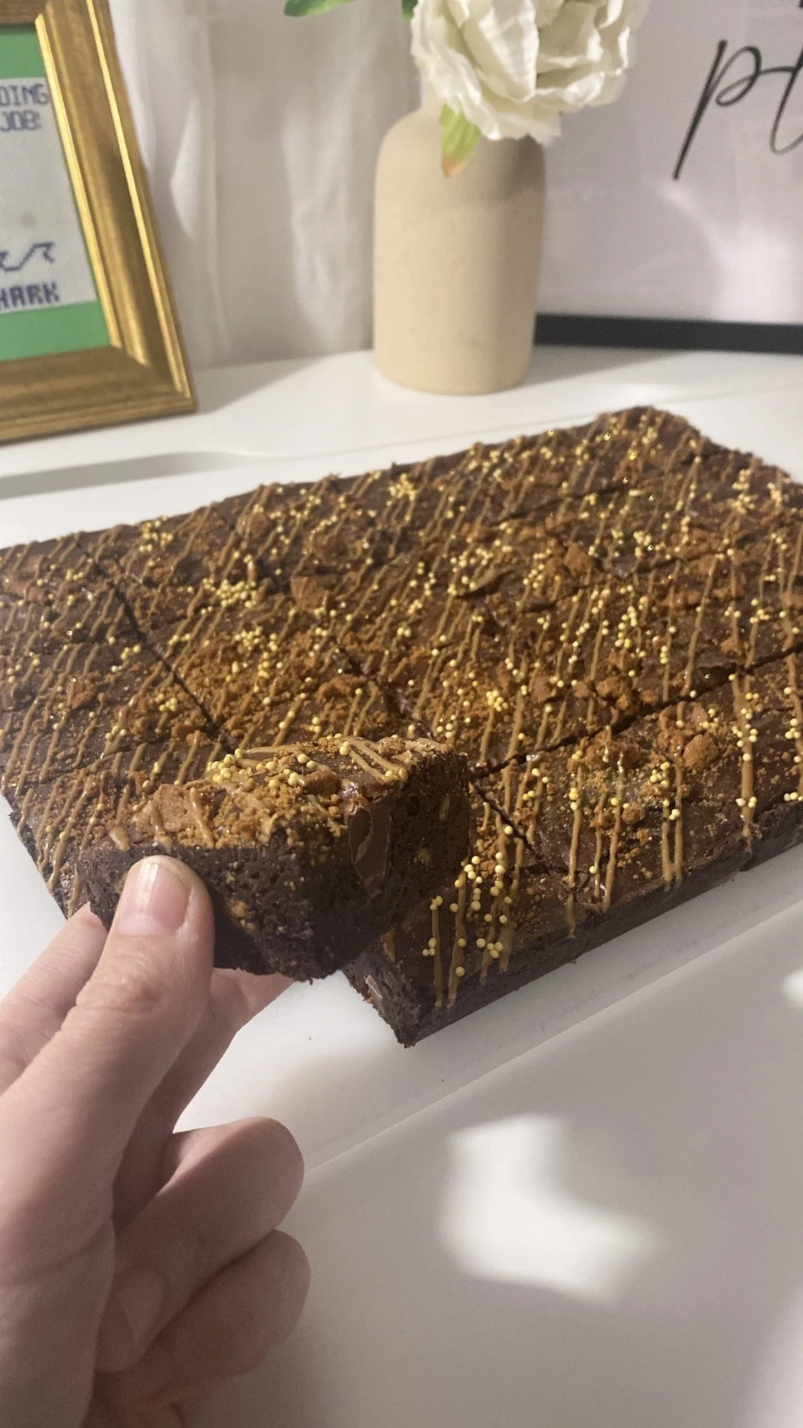 A person holding a square piece of chocolate browniewith biscoff drizzle and yellow sprinkles on top, on a white surface with a beige vase with white flowers in the background.