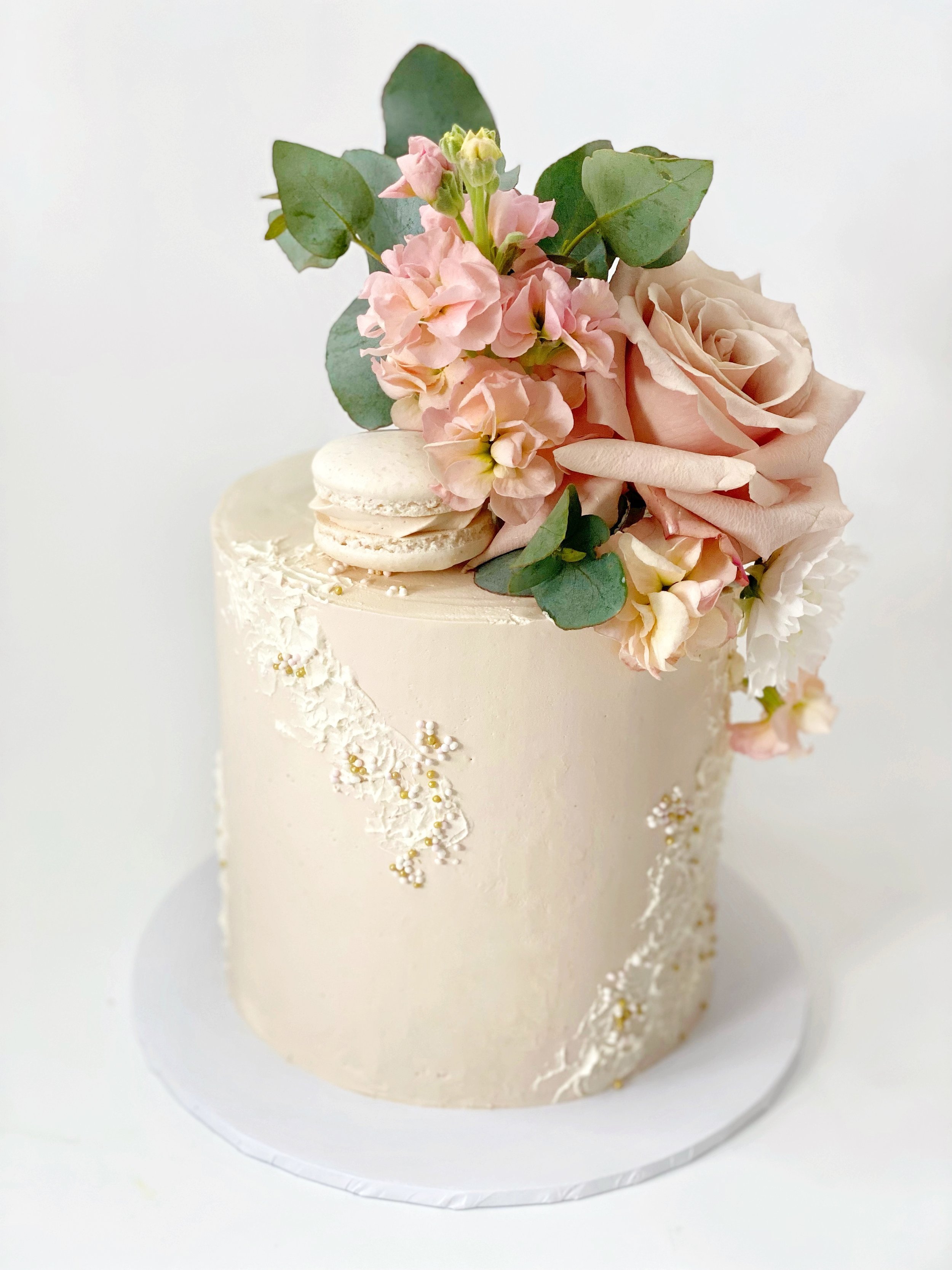 Elegant ivory wedding cake adorned with pink flowers, green leaves, and gold beads, topped with macarons.