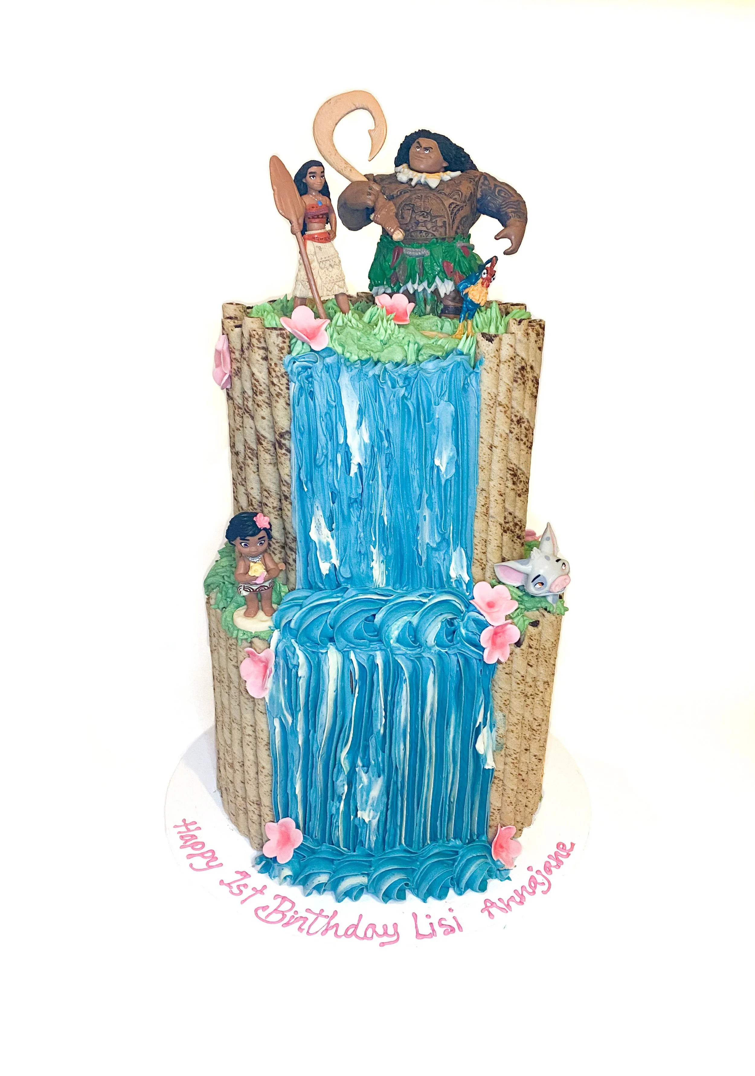A multi-tiered birthday cake themed from Moana featuring figurines of Moana, Maui, Pua, and Heihei, with blue cascading water effects and pink flowers, and a birthday message for Lisi Anne.