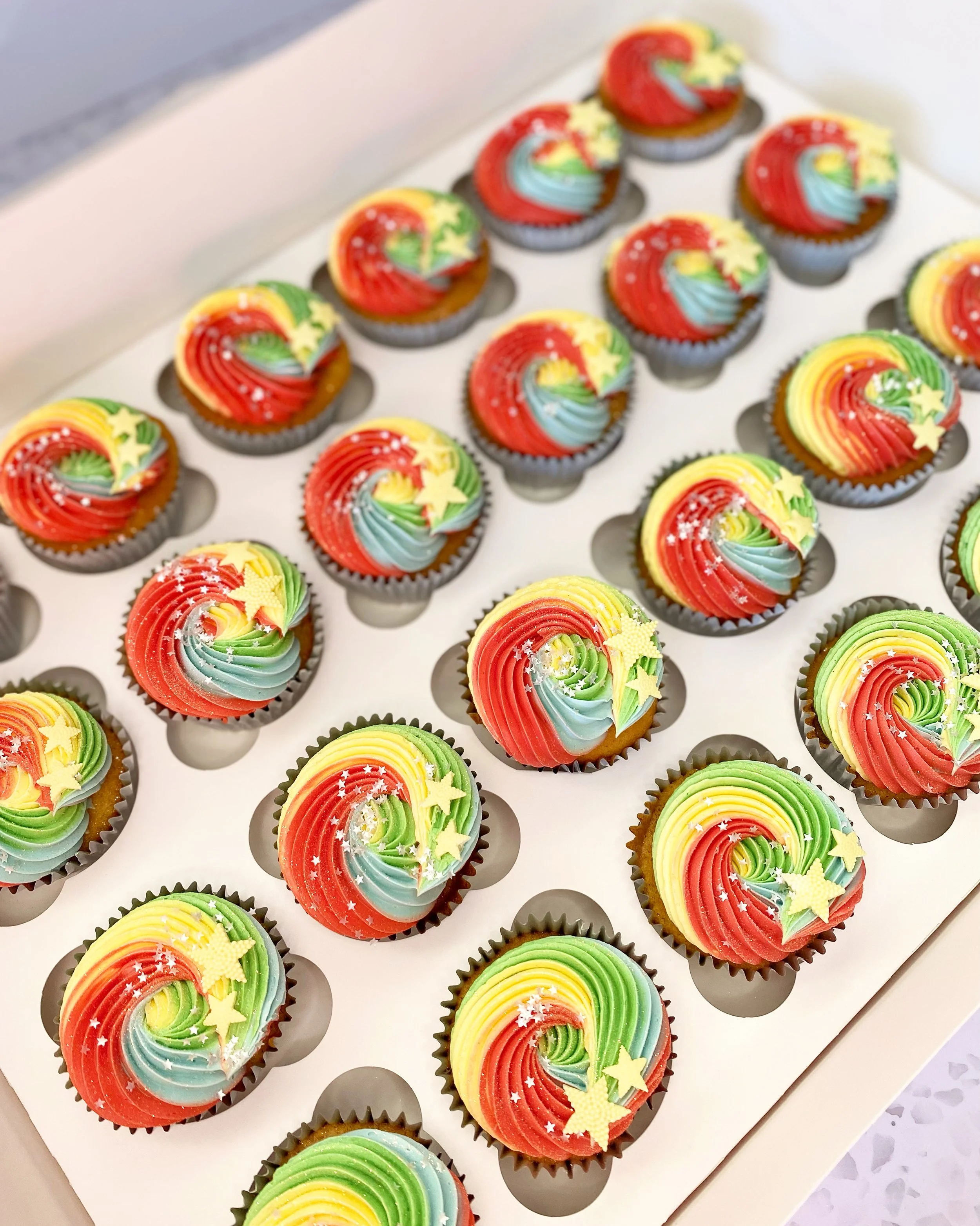 Colorful cupcakes with rainbow swirl frosting, decorated with star-shaped sprinkles and edible glitter, arranged in a white cupcake box.
