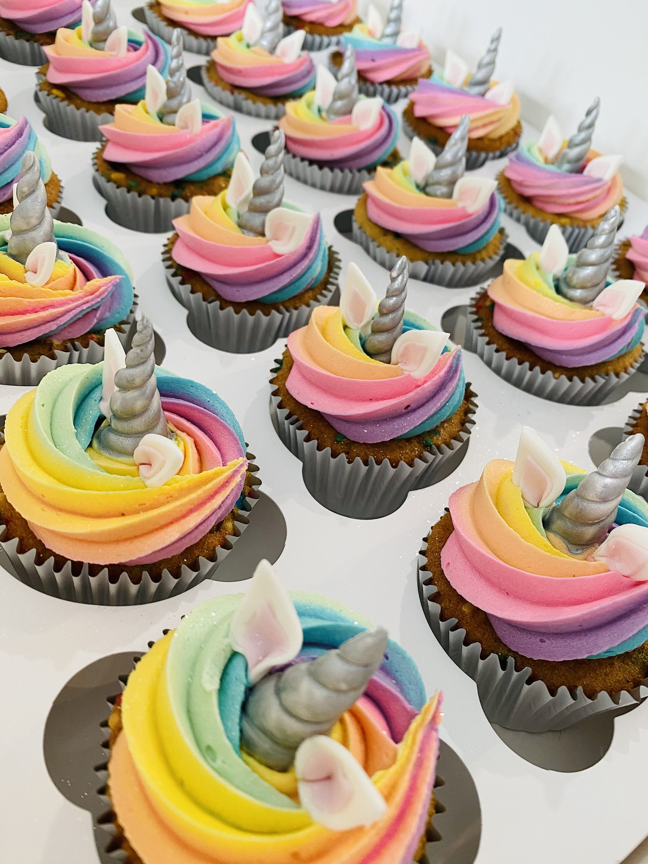 Colorful cupcakes decorated as unicorns with rainbow frosting, silver unicorn horns, and unicorn ears in cupcake wrappers.
