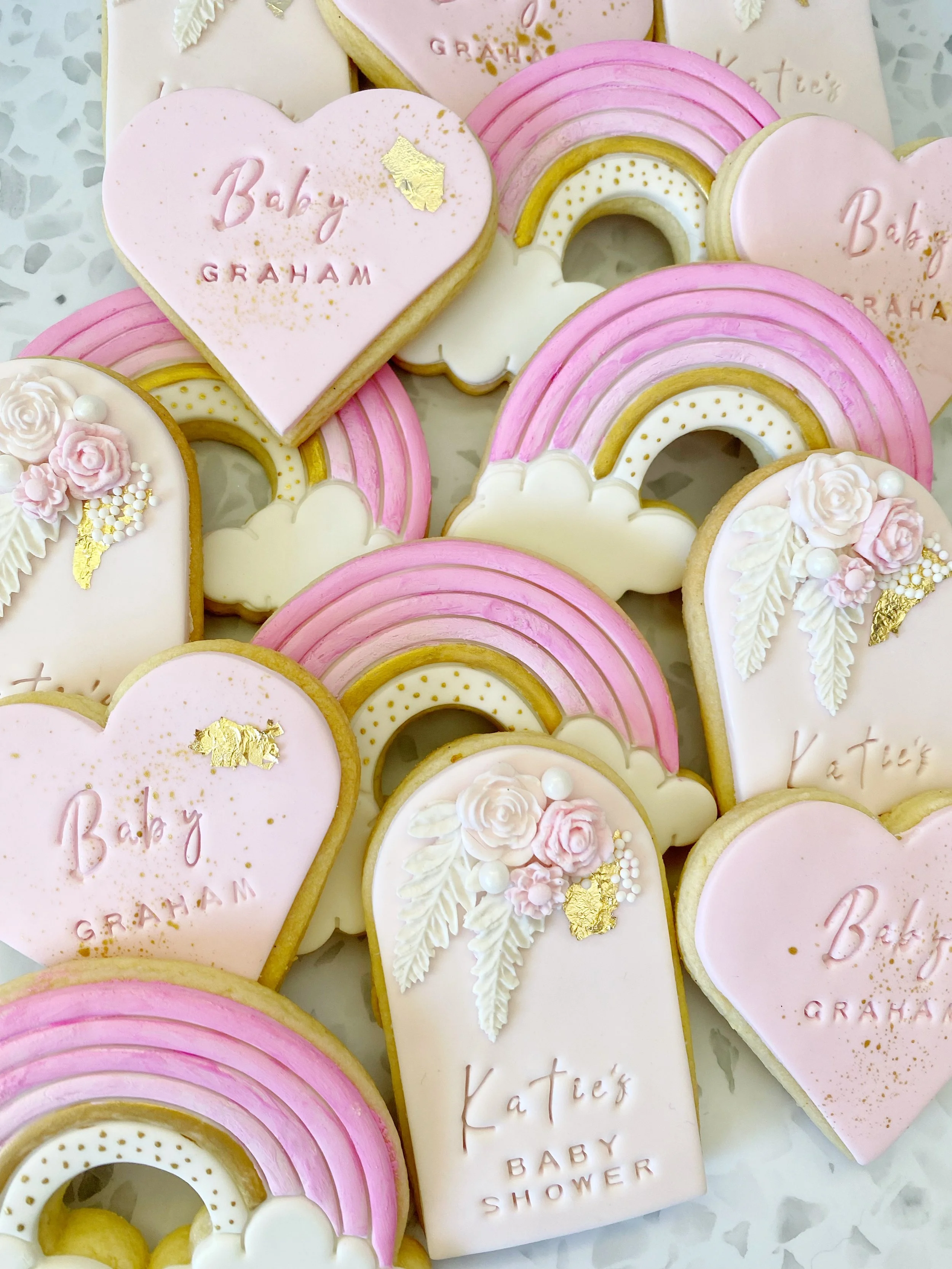 Decorative cookies with pastel pink, white, and gold icing, shaped like rainbows, hearts, and clouds. Some cookies have inscriptions like 'Baby Graham' and 'Katelyn Baby Shower,' with floral and gold accents.