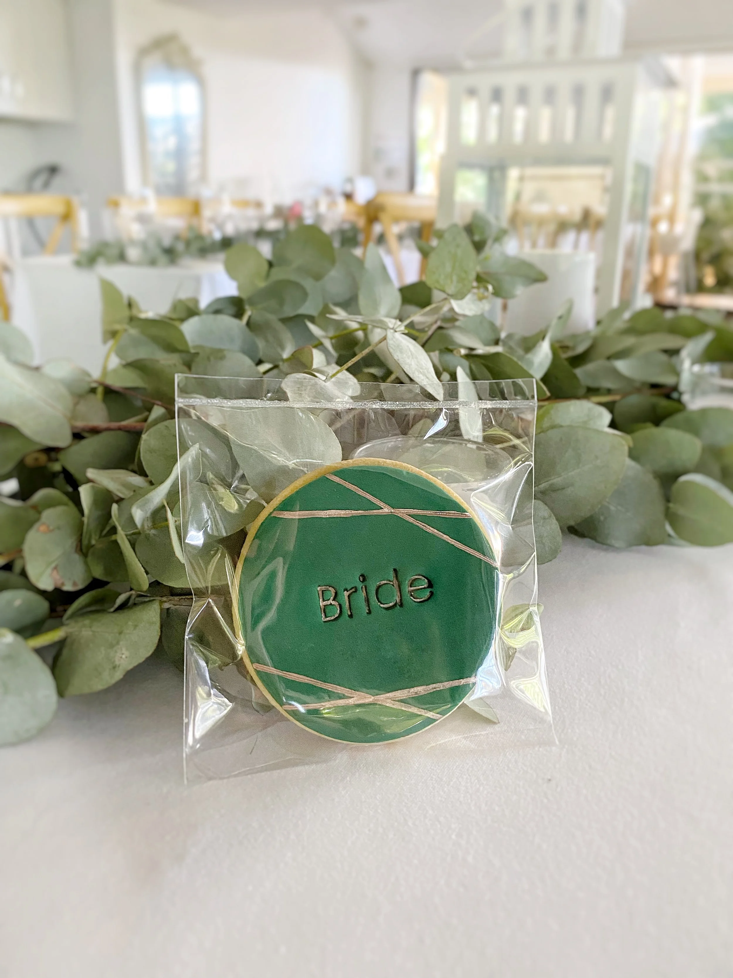 A green cookie with the word "Bride" written on it, wrapped in clear plastic, sits on a white surface with green foliage in the background, at a wedding reception.