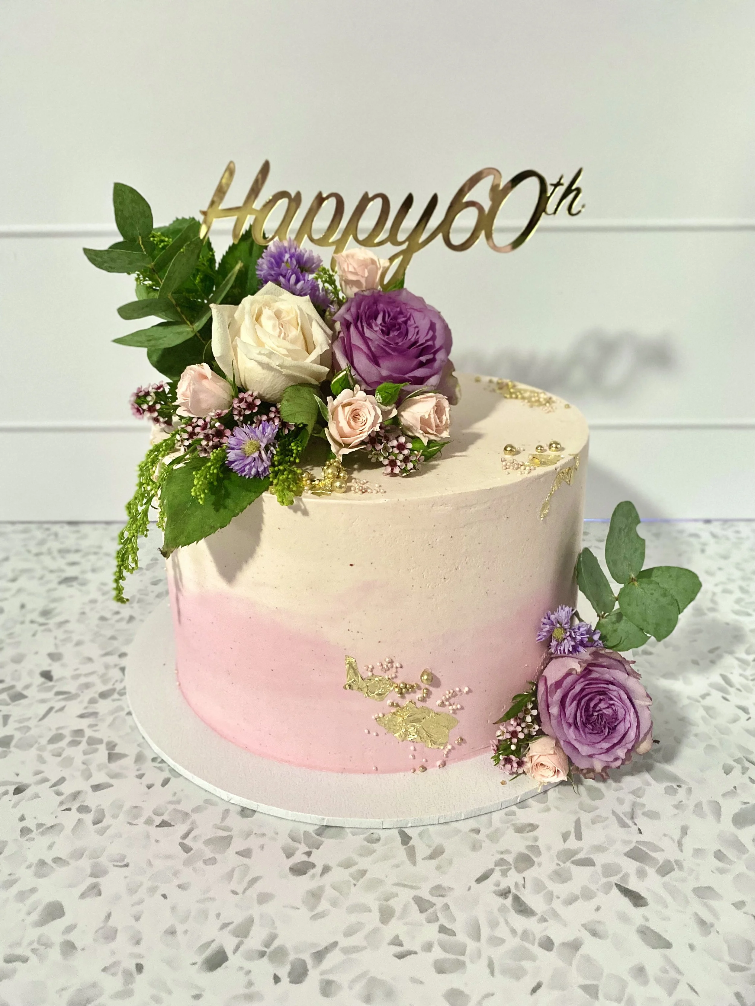 A pink and white gradient birthday cake decorated with pink and purple roses, purple flowers, greenery, gold accents, and a gold "Happy 60th" topper, placed on a white speckled surface.