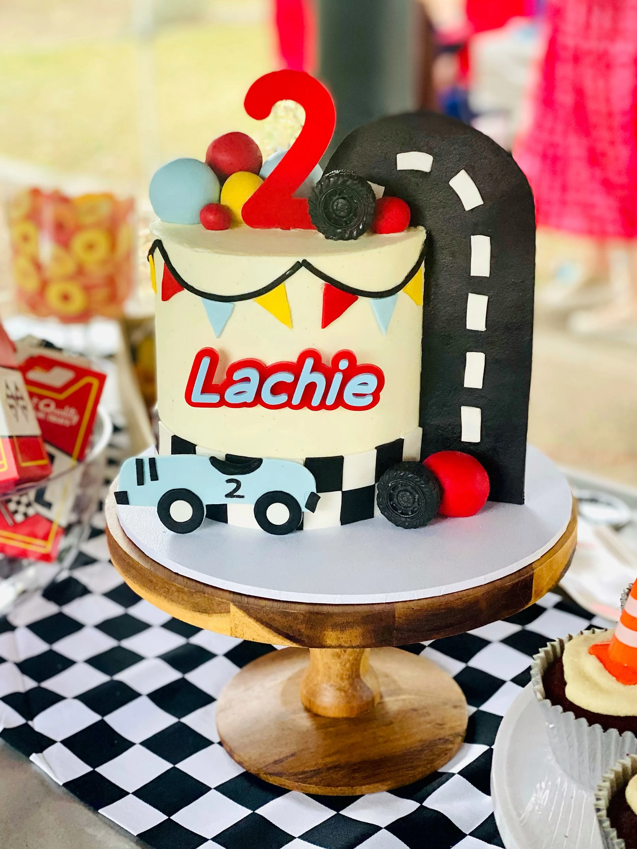 A race car-themed birthday cake with a checkered pattern and racing decorations, featuring the name 'Lachie' and the number 2, topped with colorful balls, a red number 2, and racing-inspired fondant embellishments.