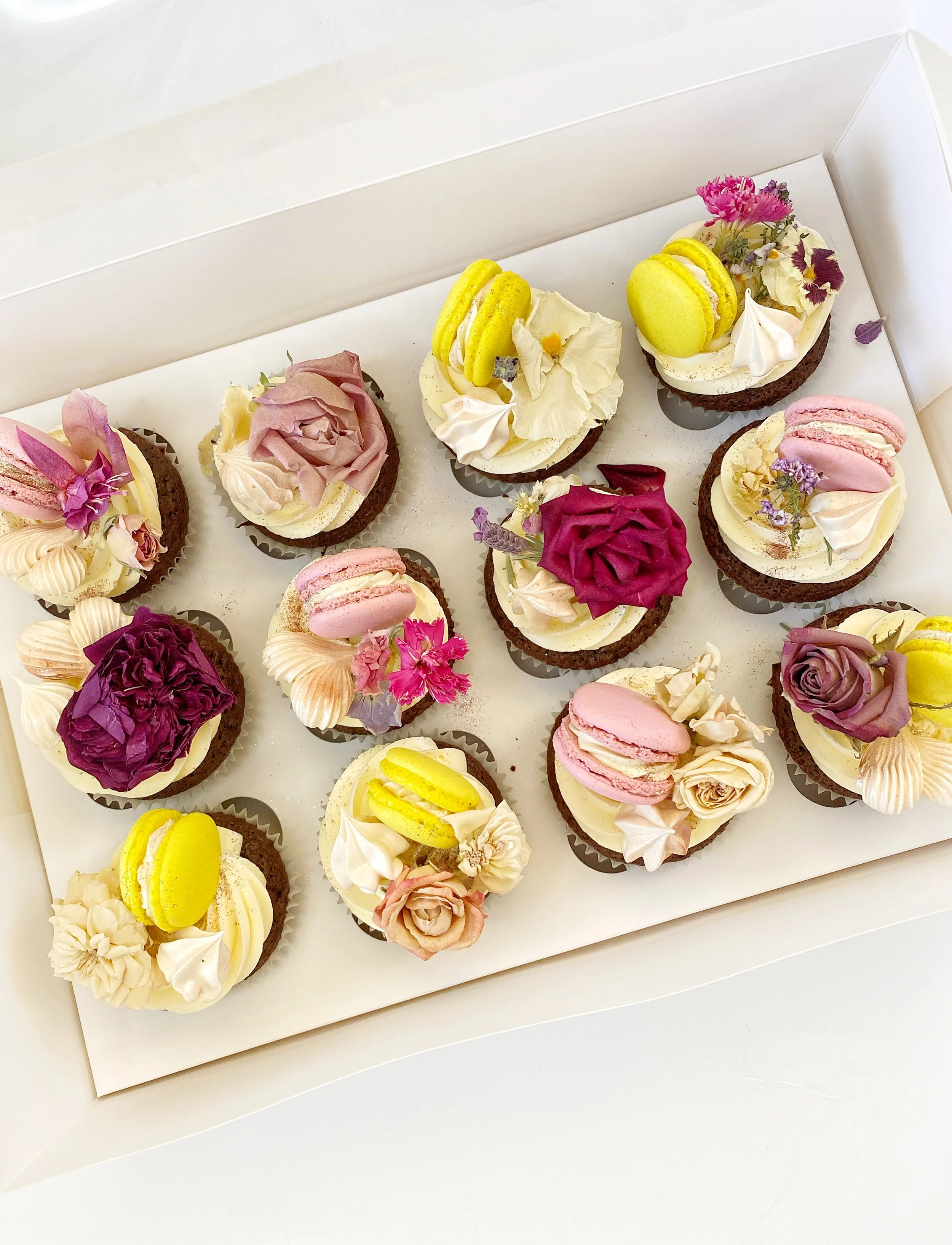 A box of decorated cupcakes with flowers, macarons, and frosting in pastel colors.