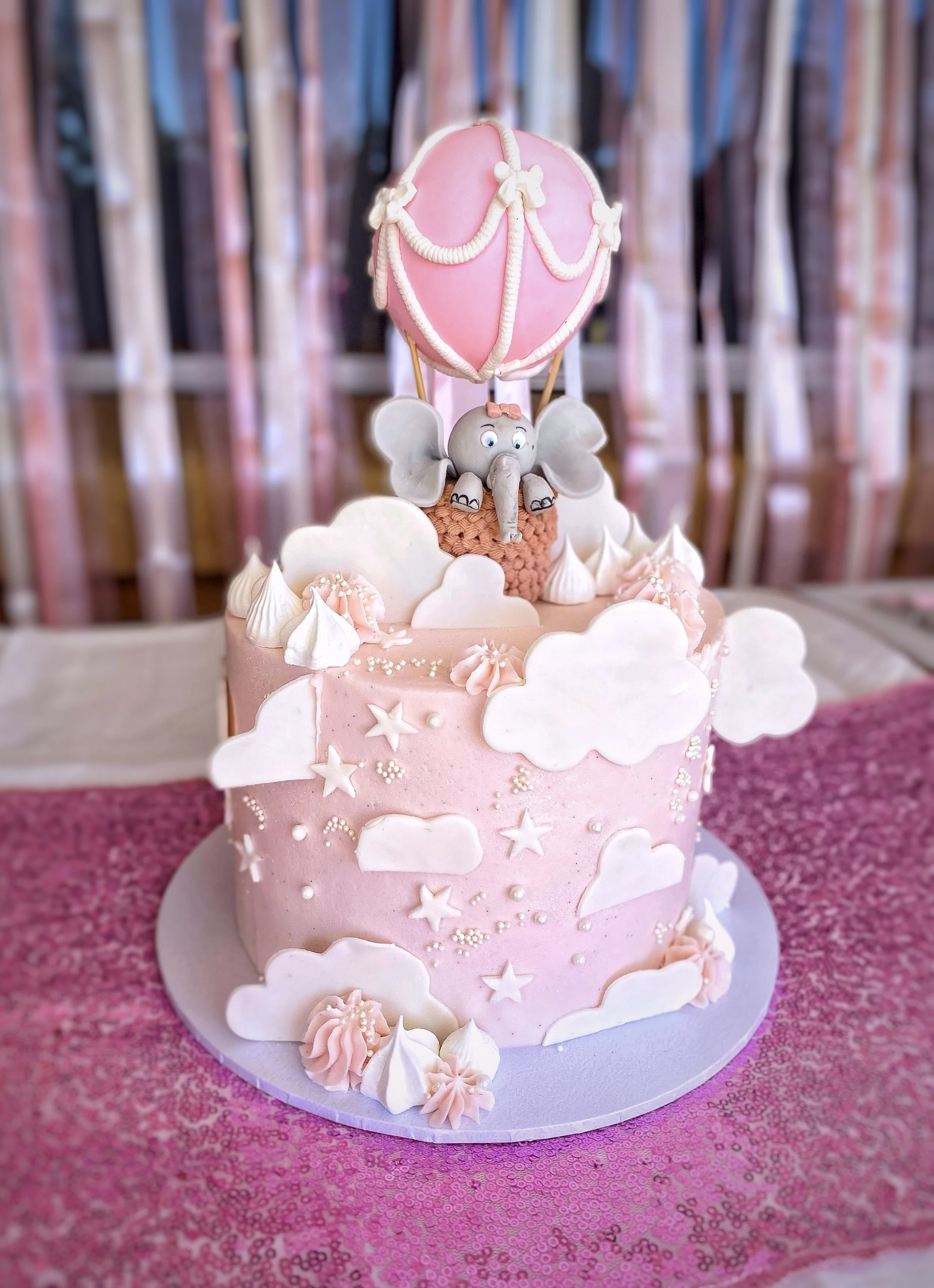 Pink birthday cake decorated with white clouds, stars, and small pearls, featuring a fondant elephant with large ears and a pink bow, riding in a basket under a pink hot air balloon with white rope details.