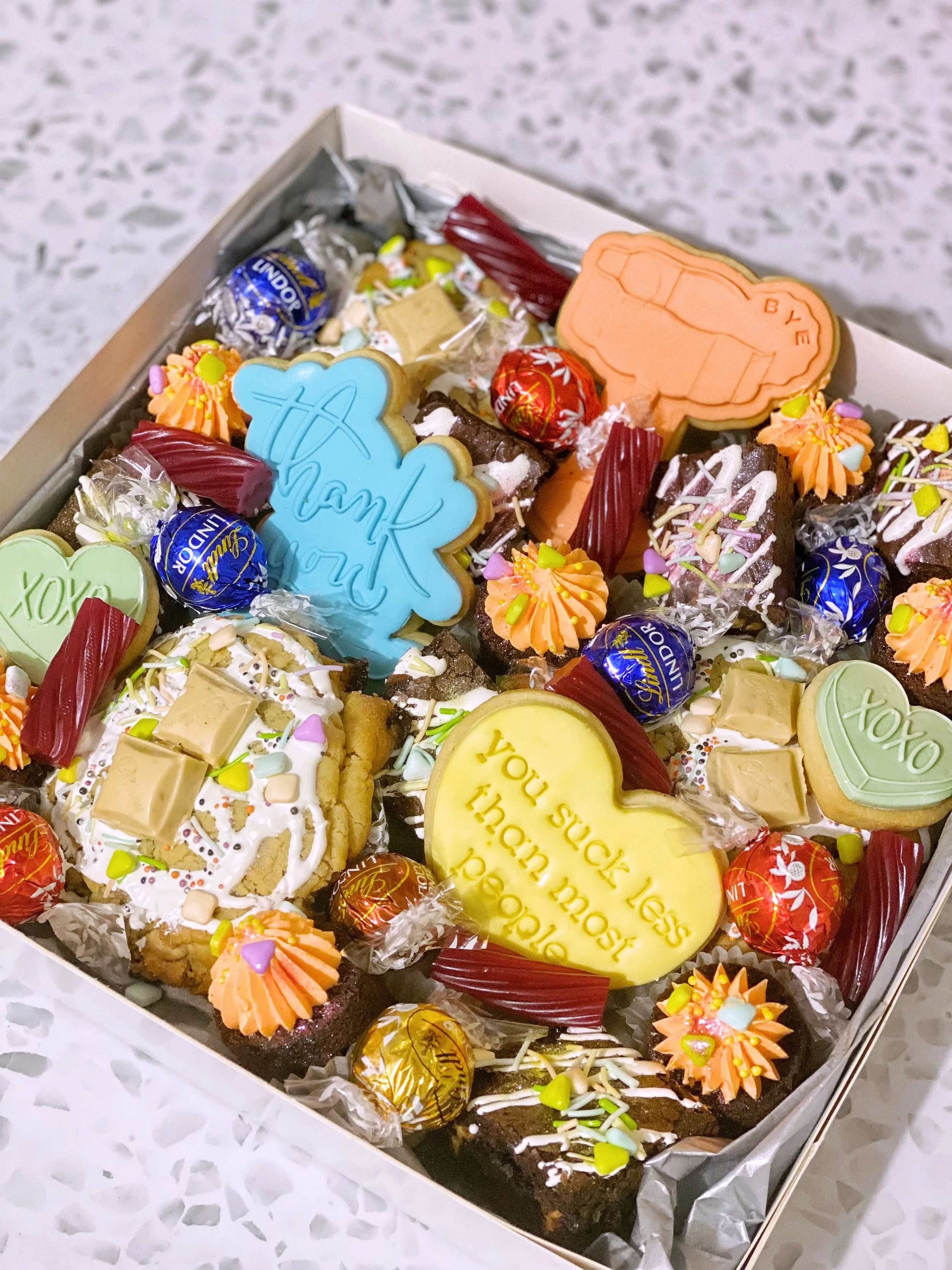 Box of assorted decorated cookies and chocolates including heart-shaped, cloud-shaped, and rectangular cookies with messages, surrounded by wrapped chocolates and candies.