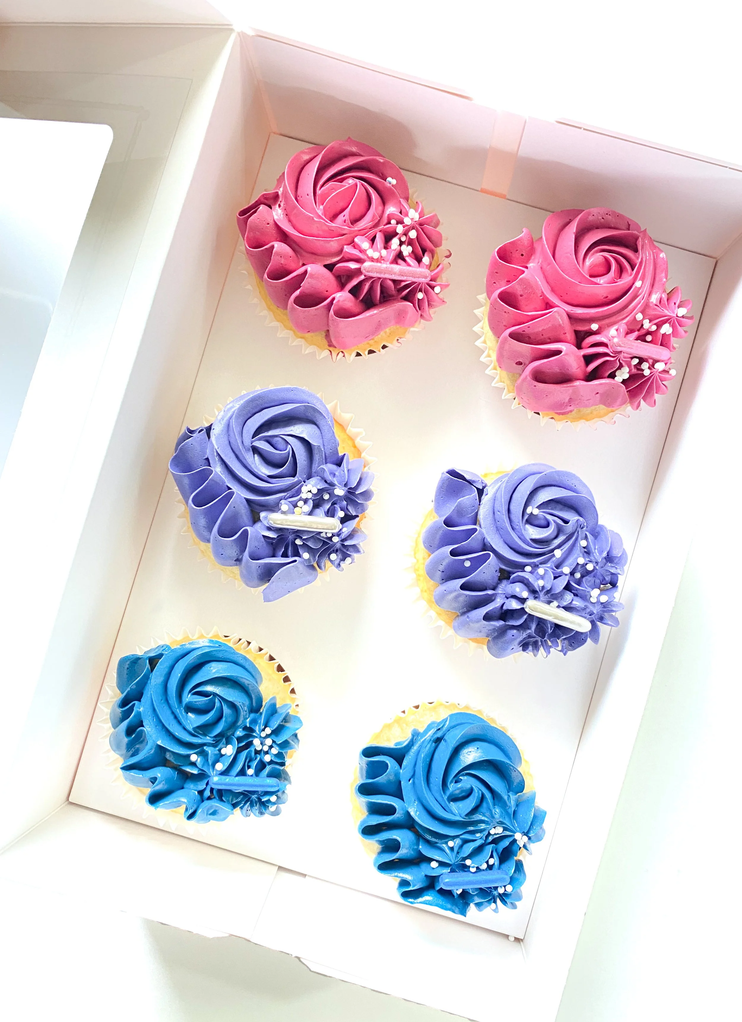 Six decorated cupcakes with buttercream swirls in pink, purple, and blue with sprinkles, inside a white box.