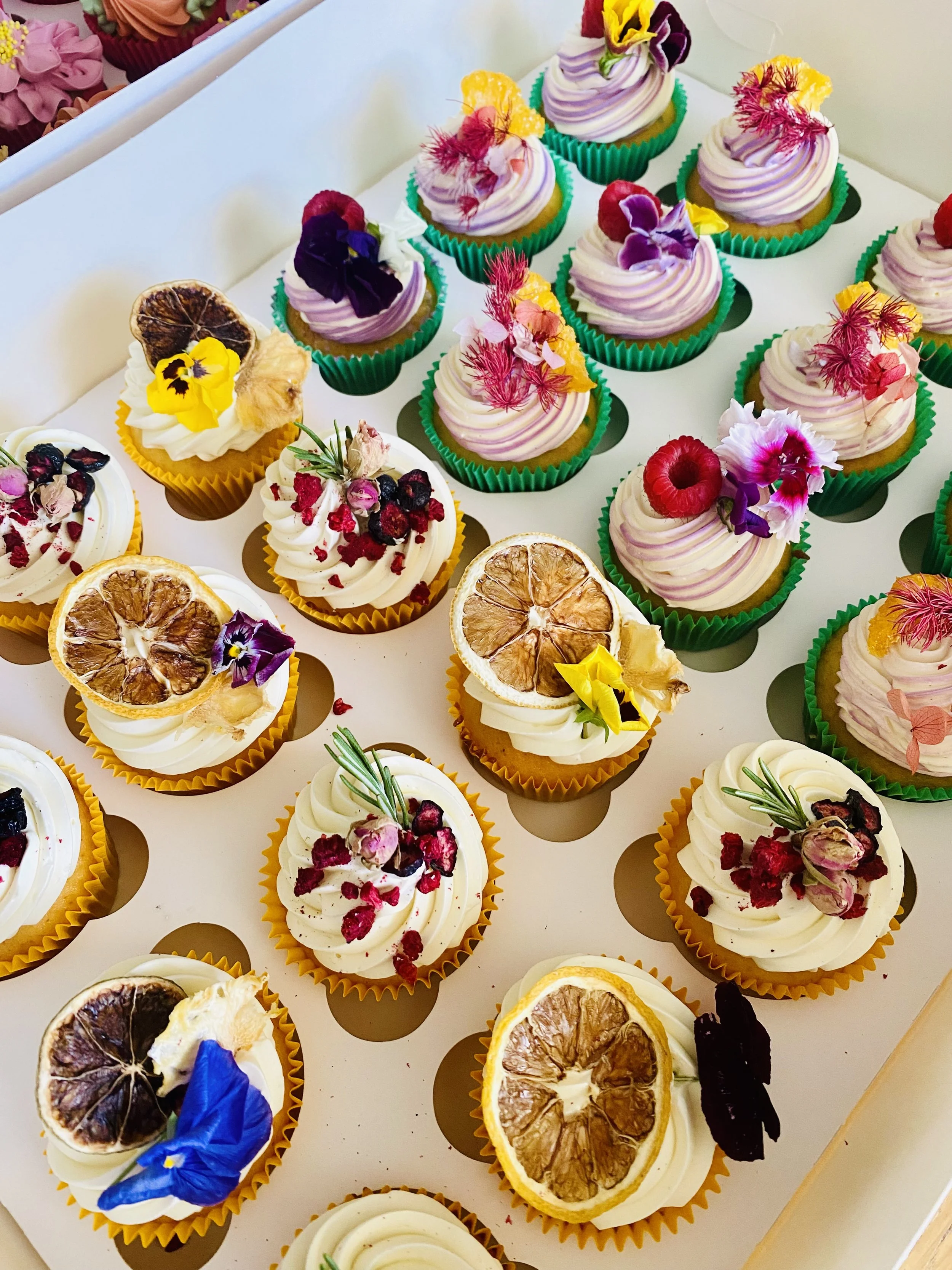 Assorted cupcakes with frosting and edible decorations such as dried citrus slices, berries, flowers, and herbs arranged in colorful cupcake liners.