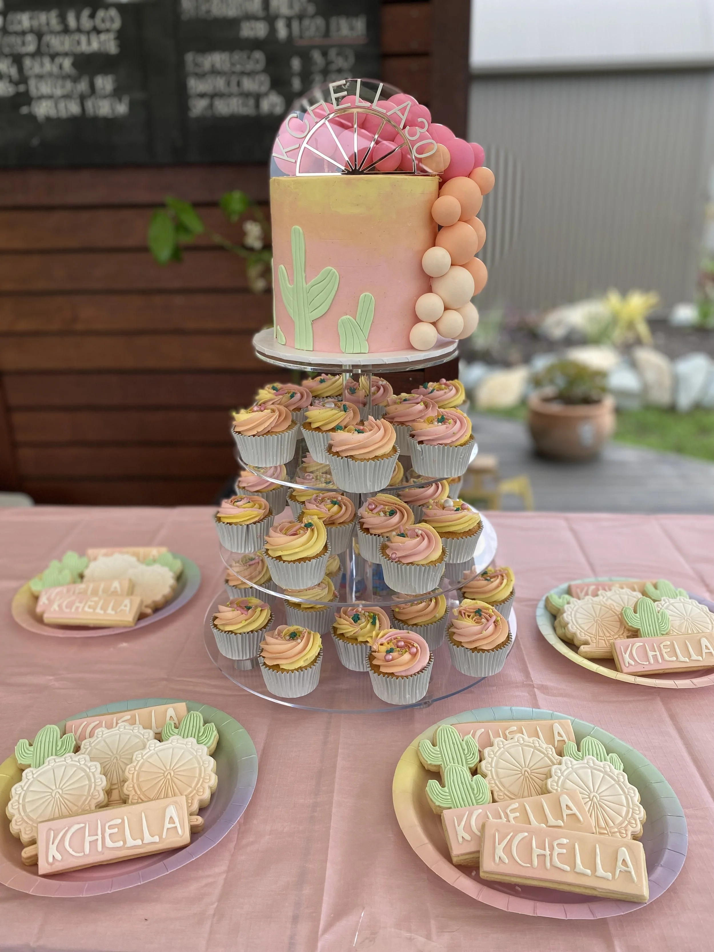 A pink birthday cake with cactus decorations and a balloon topper reading "KCHELLA 30" on a tiered stand. Surrounding cupcakes with pink and yellow swirled frosting. Plates with cactus-shaped cookies and rectangular cookies with the name "KCHELLA" wr