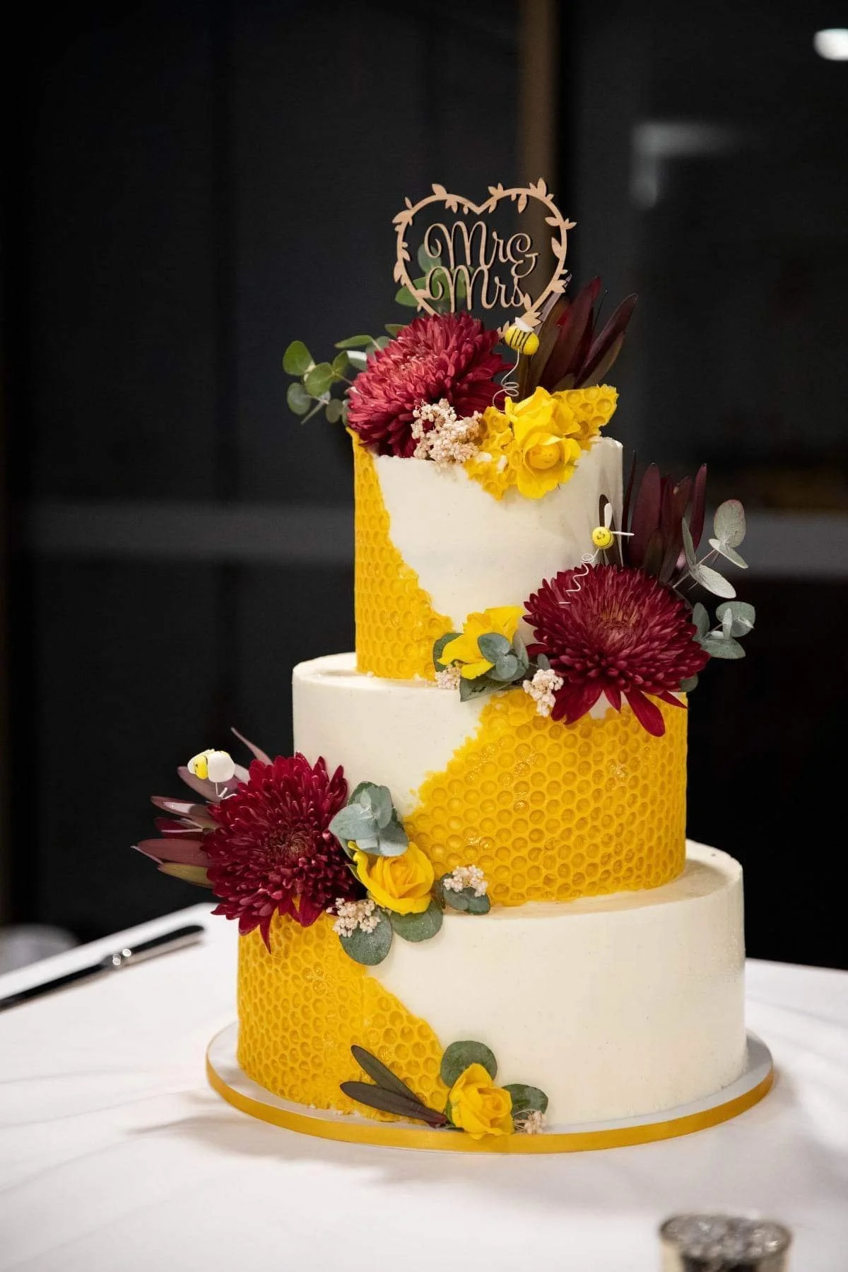 A three-tier wedding cake decorated with yellow honeycomb pattern, large red flowers, green leaves, and a "Mrs & Mrs" topper.