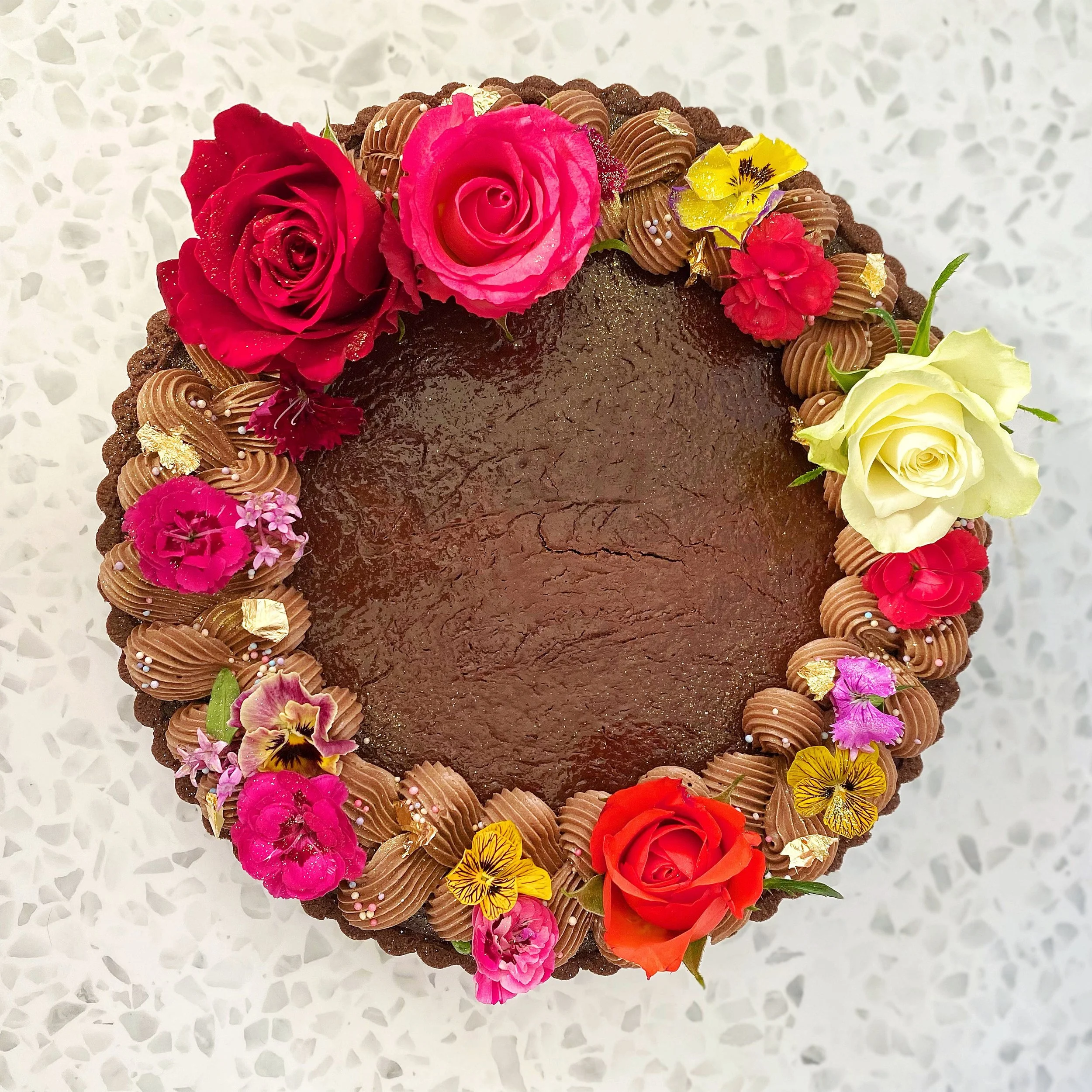 Round chocolate tartdecorated with colorful fresh flowers including red, yellow, pink, and white roses, and various smaller flowers, with piped chocolate chantilly around the edges, on a white speckled surface.