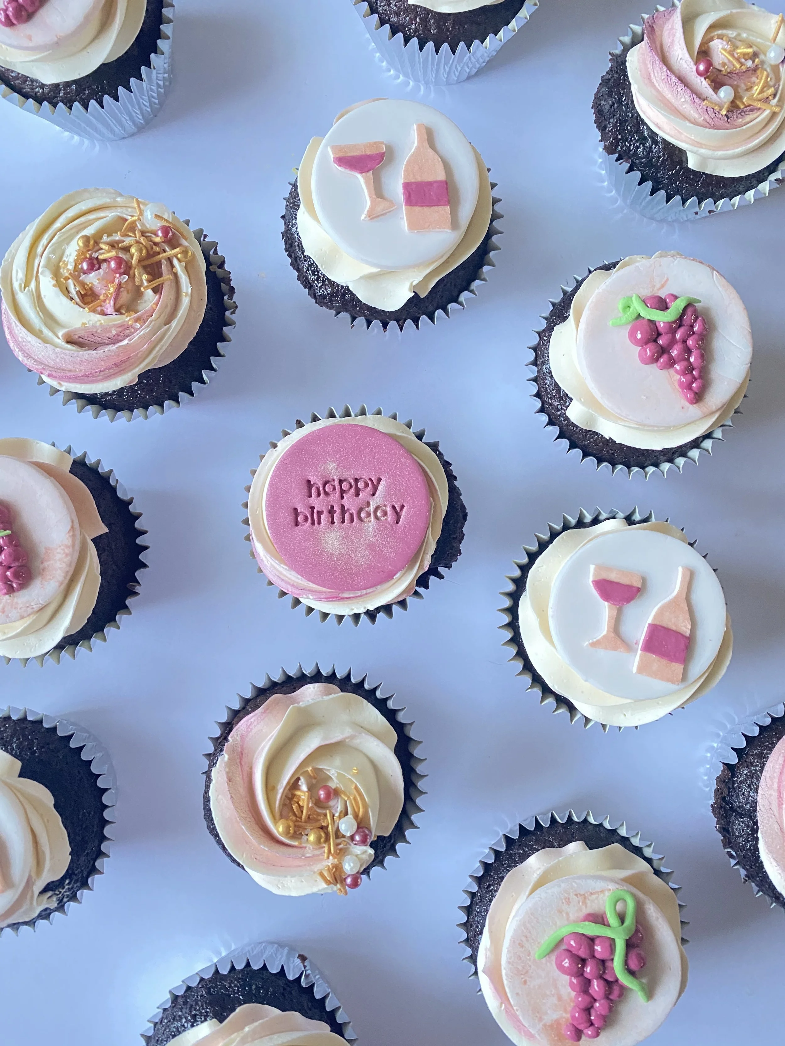 Assorted cupcakes with pink, white, and gold decorations, including one with a 'happy birthday' message, and others with images of wine bottles, glasses, and grape clusters.