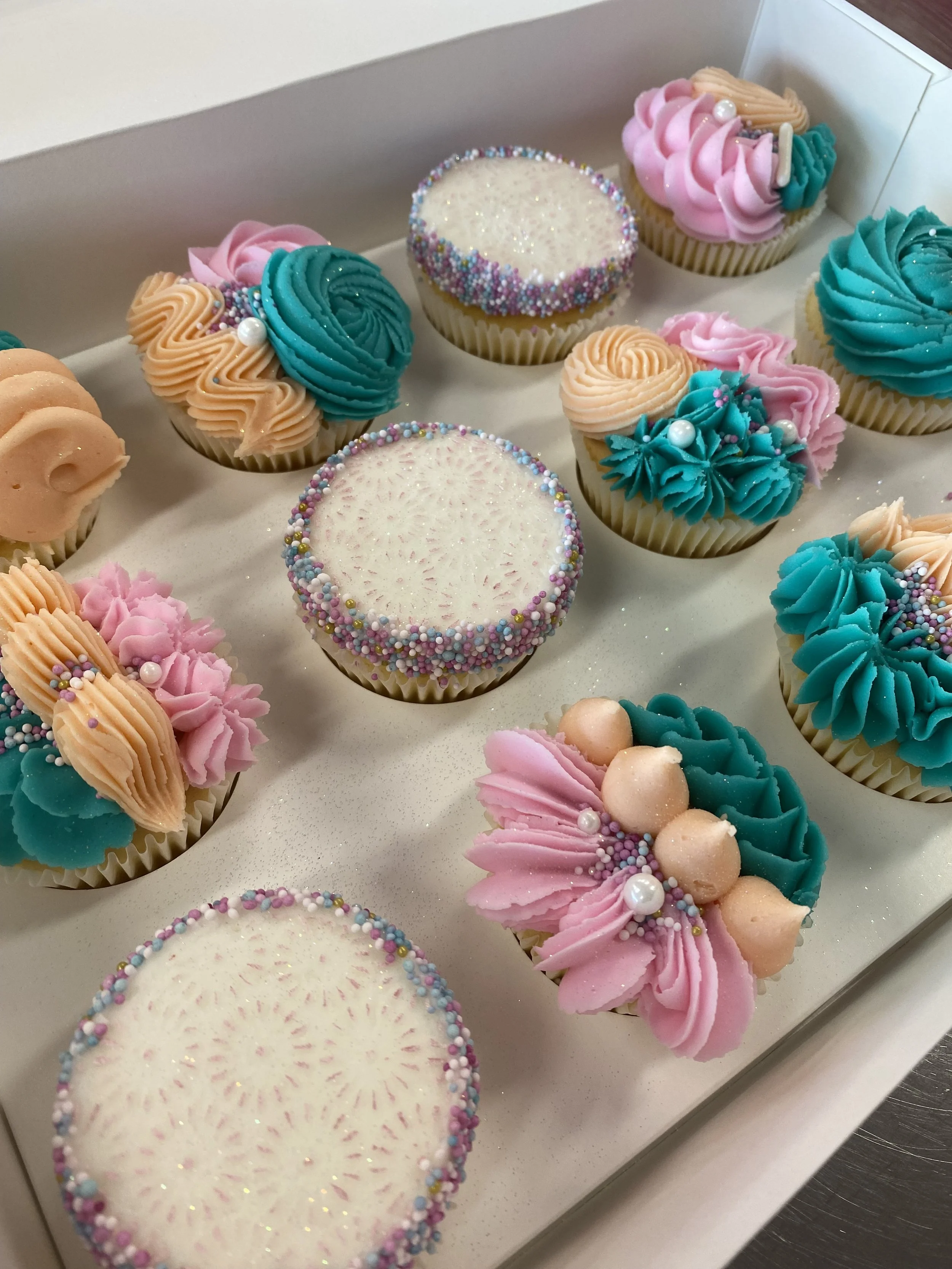A box of decorated cupcakes with pastel-colored frosting, sprinkles, and pearl toppings.