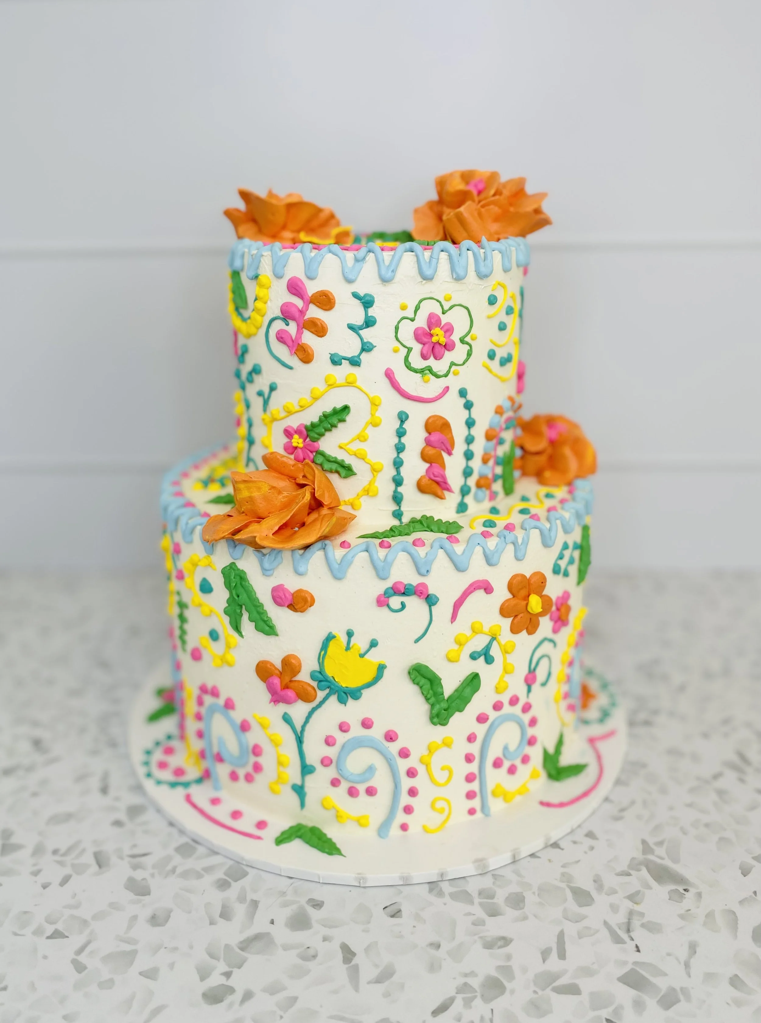 Colorfully decorated two-tier birthday cake with floral and abstract icing patterns, topped with orange edible flowers.