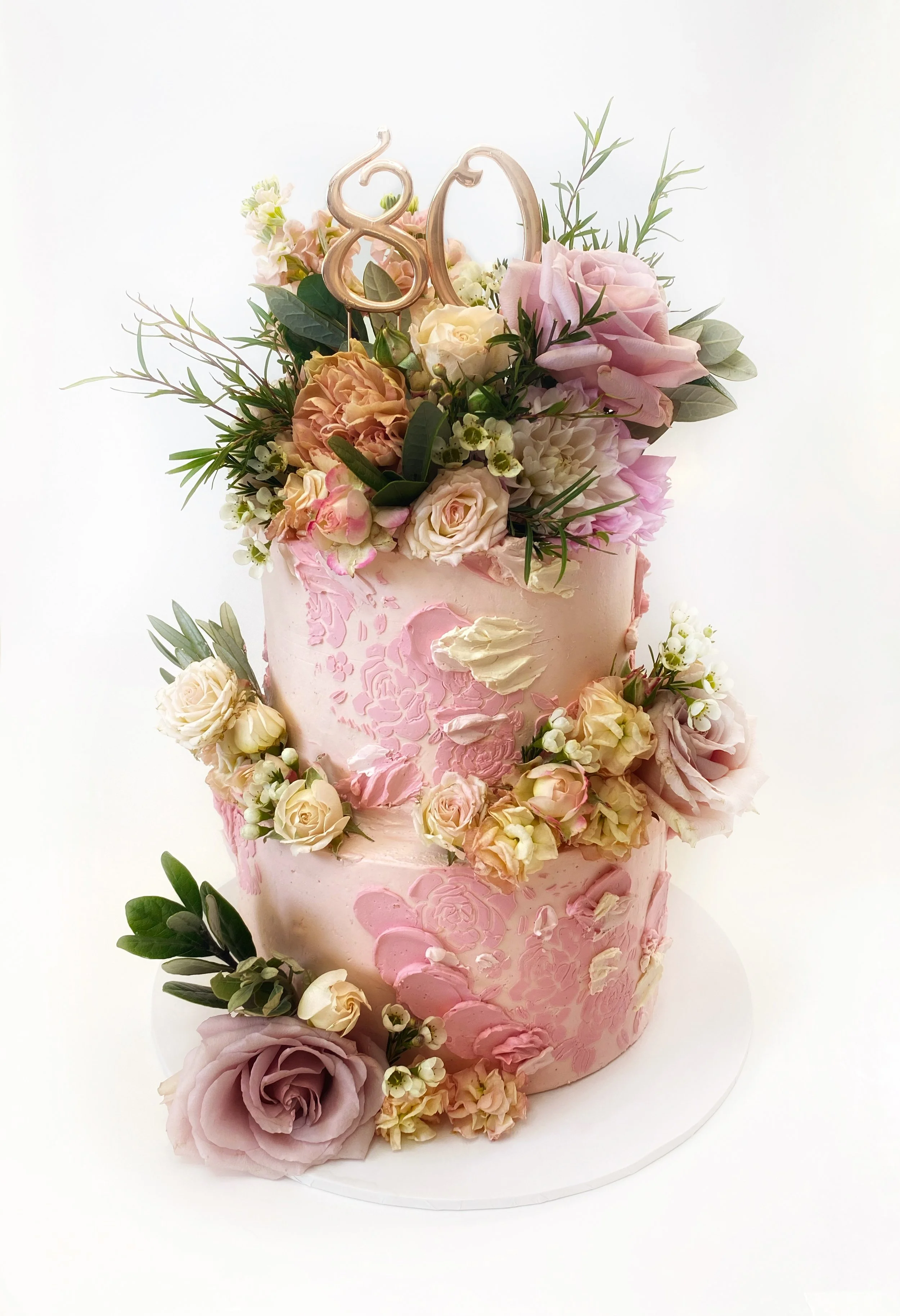 Two-tier pink floral cake decorated with roses, carnations, greenery, and a gold '80' cake topper.