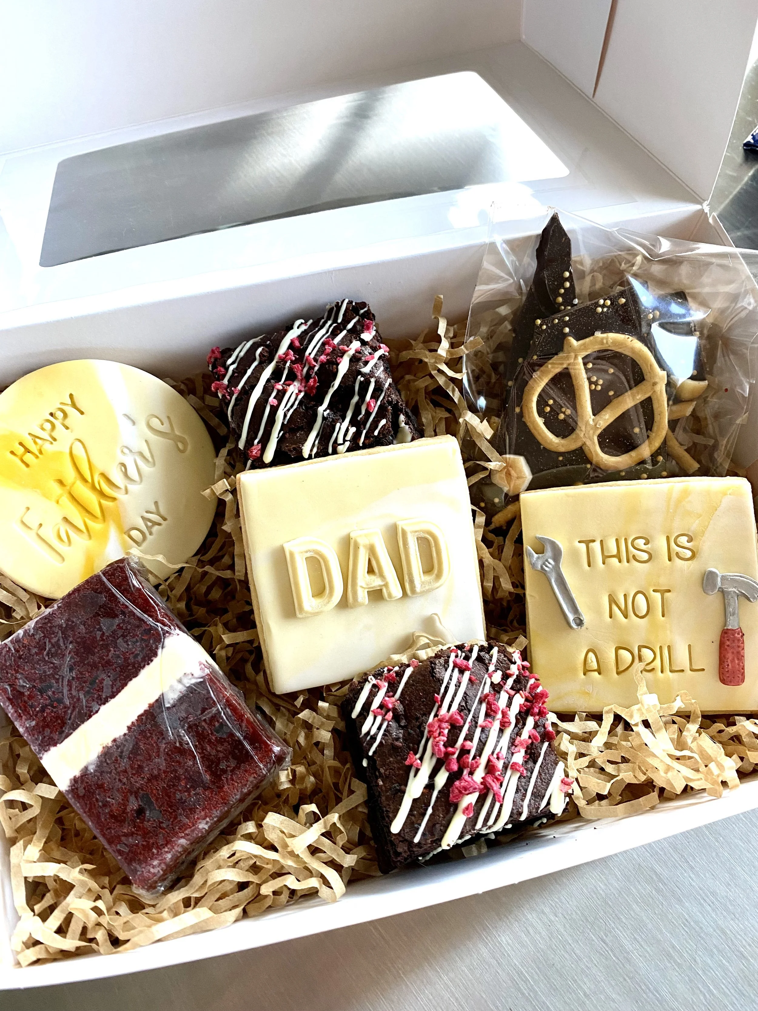 A gift box with assorted decorated chocolates and brownies, including one with the message 'Happy Father's Day', one with 'DAD' on white chocolate, and another with 'This is not a drill' and tools graphics.