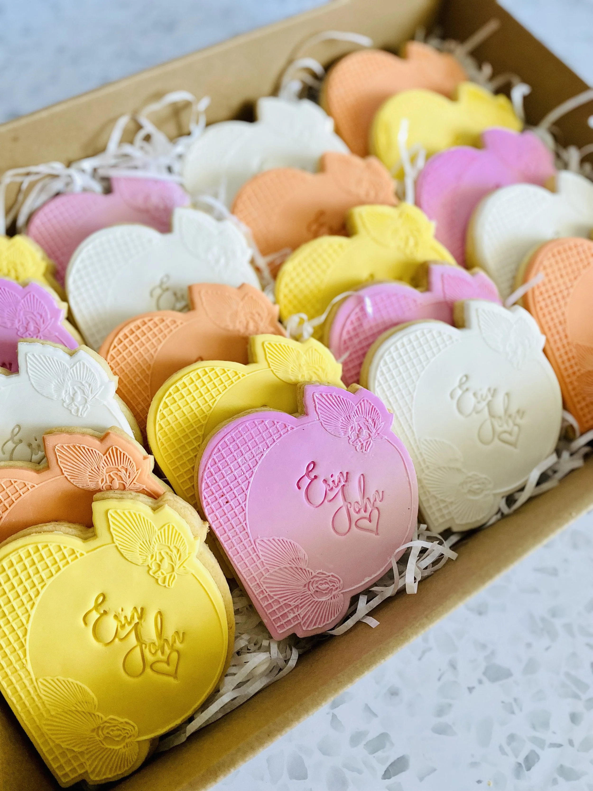 Assorted pastel-colored cookies with 'Eat' and 'Love' written on them, arranged in a cardboard box with shredded paper filling.